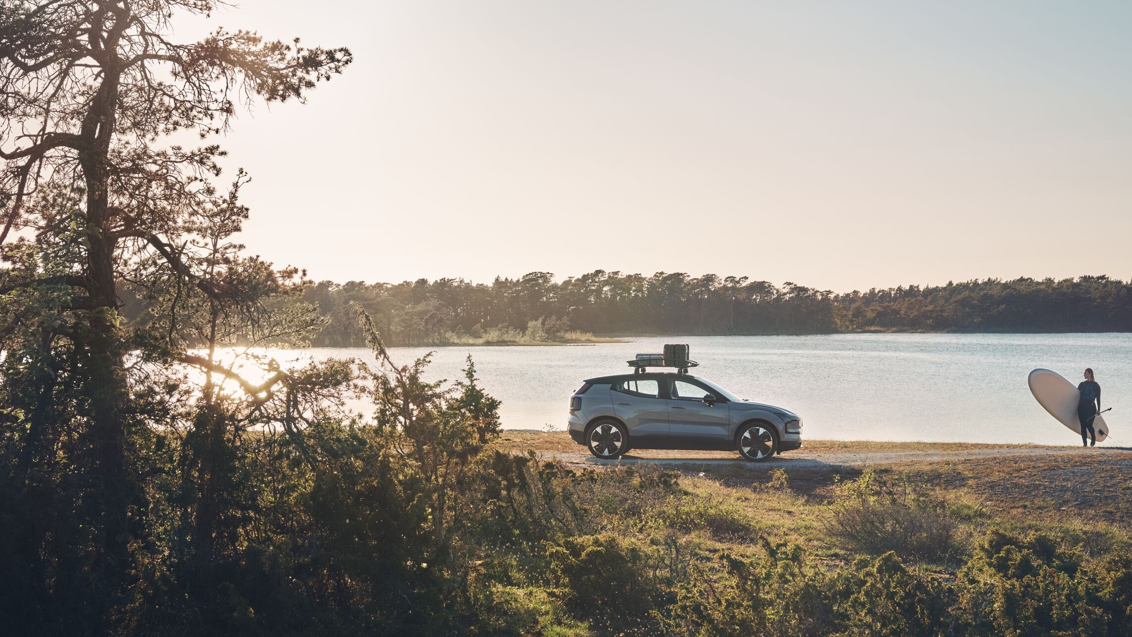 Seitenansicht des Volvo EX30 mit einer Frau, die ein Surferbrett neben dem Wasser hält.