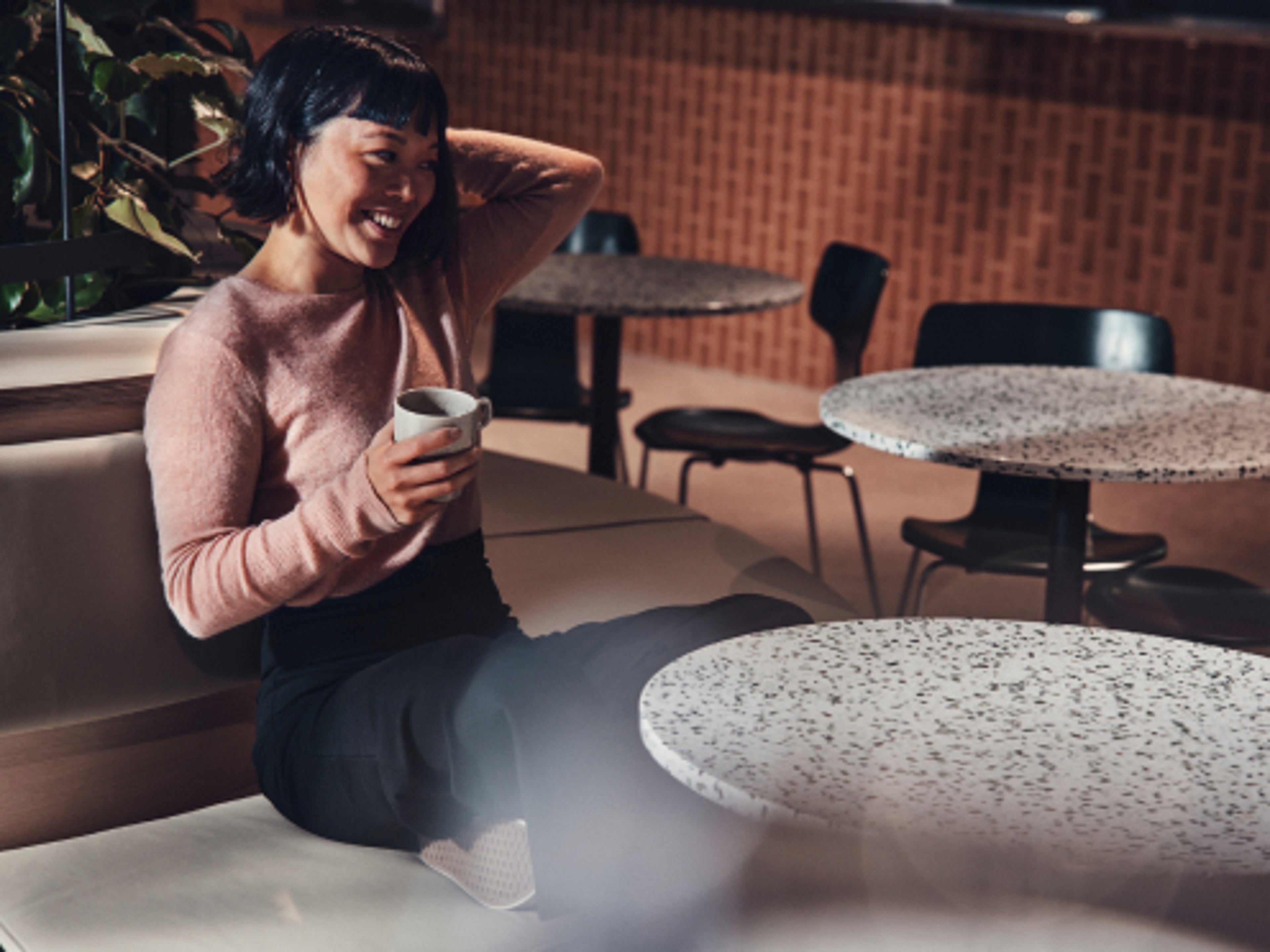A woman sat with a cup by a table smiling at something.