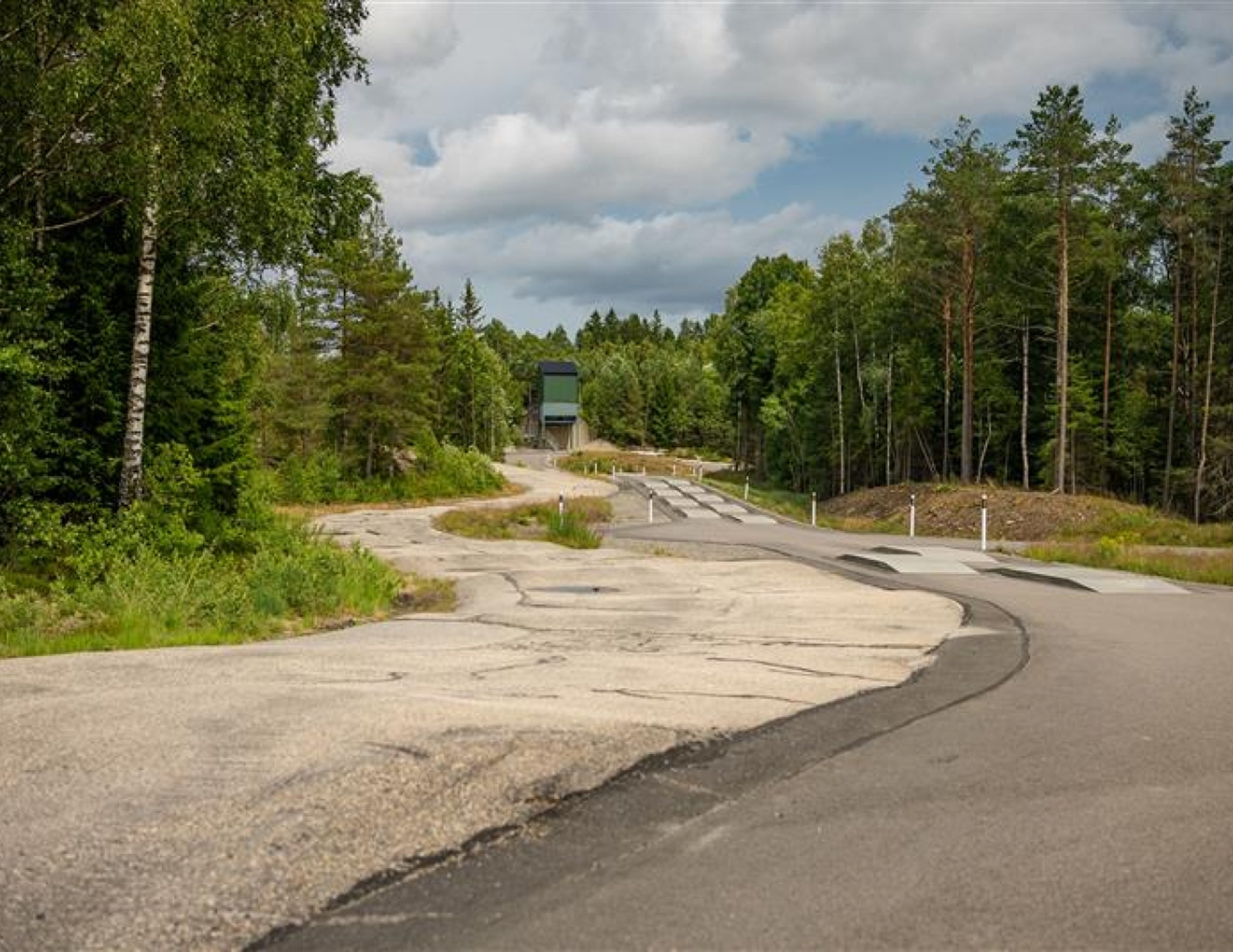 En slingrande väg omgiven av täta gröna träd och molnig himmel, med en liten byggnad synlig i fjärran.