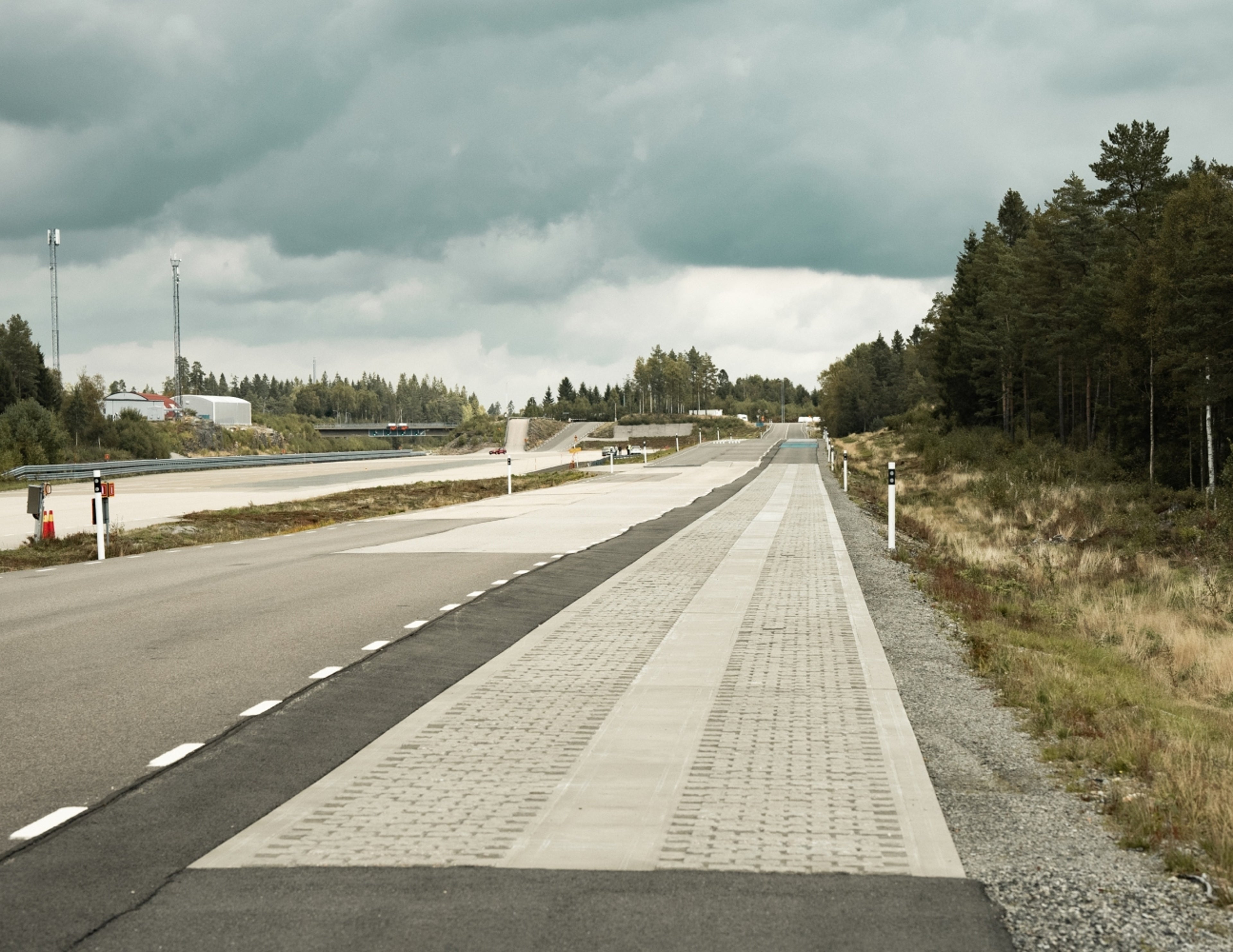 En bred, tom testbana sträcker sig i fjärran under en molnig himmel, kantad av träd och gräsytor, utan fordon eller människor synliga på vägen.
