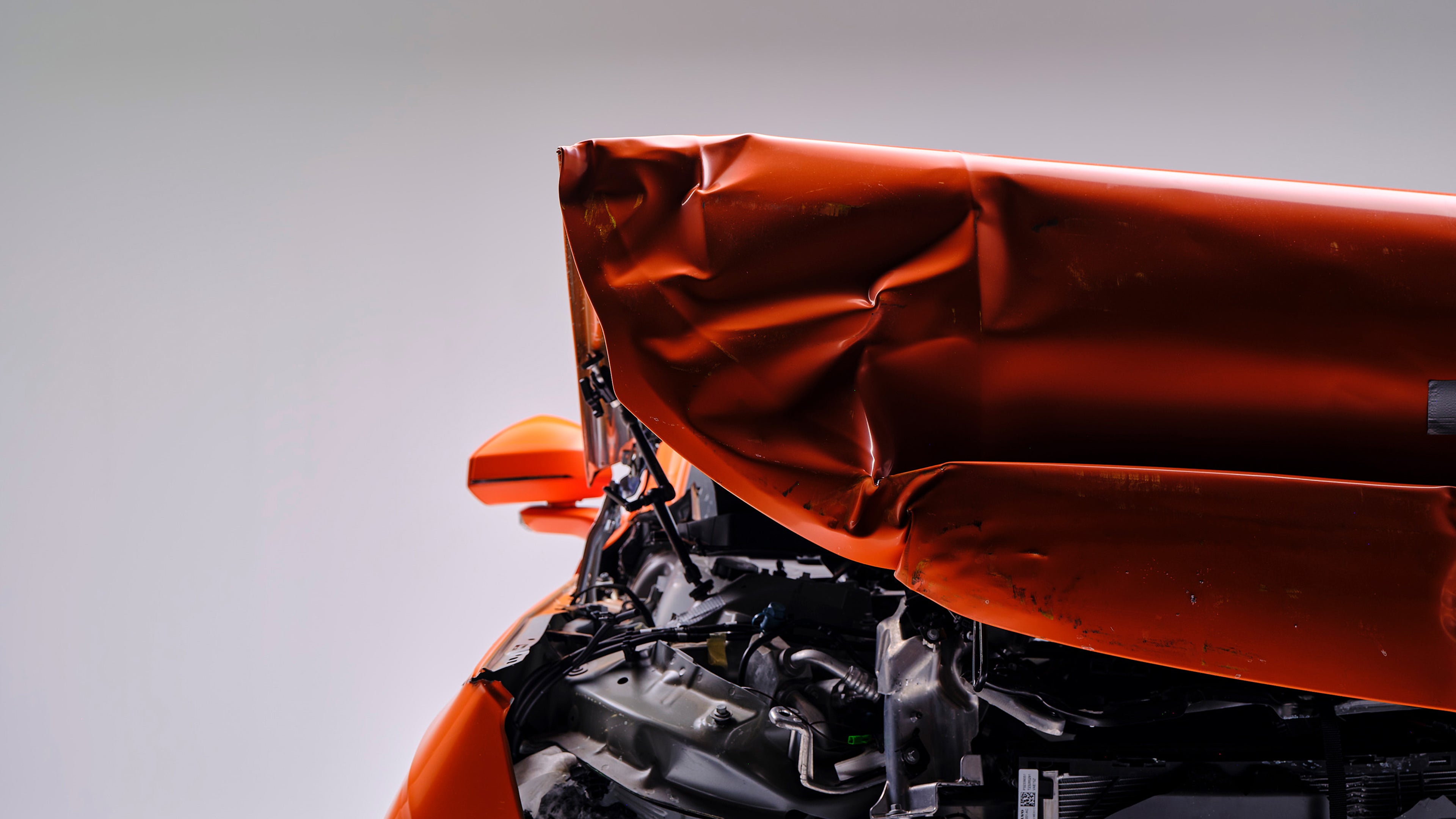 Vista frontale di un’auto arancio sottoposta a crash test con cofano accartocciato e motore esposto.