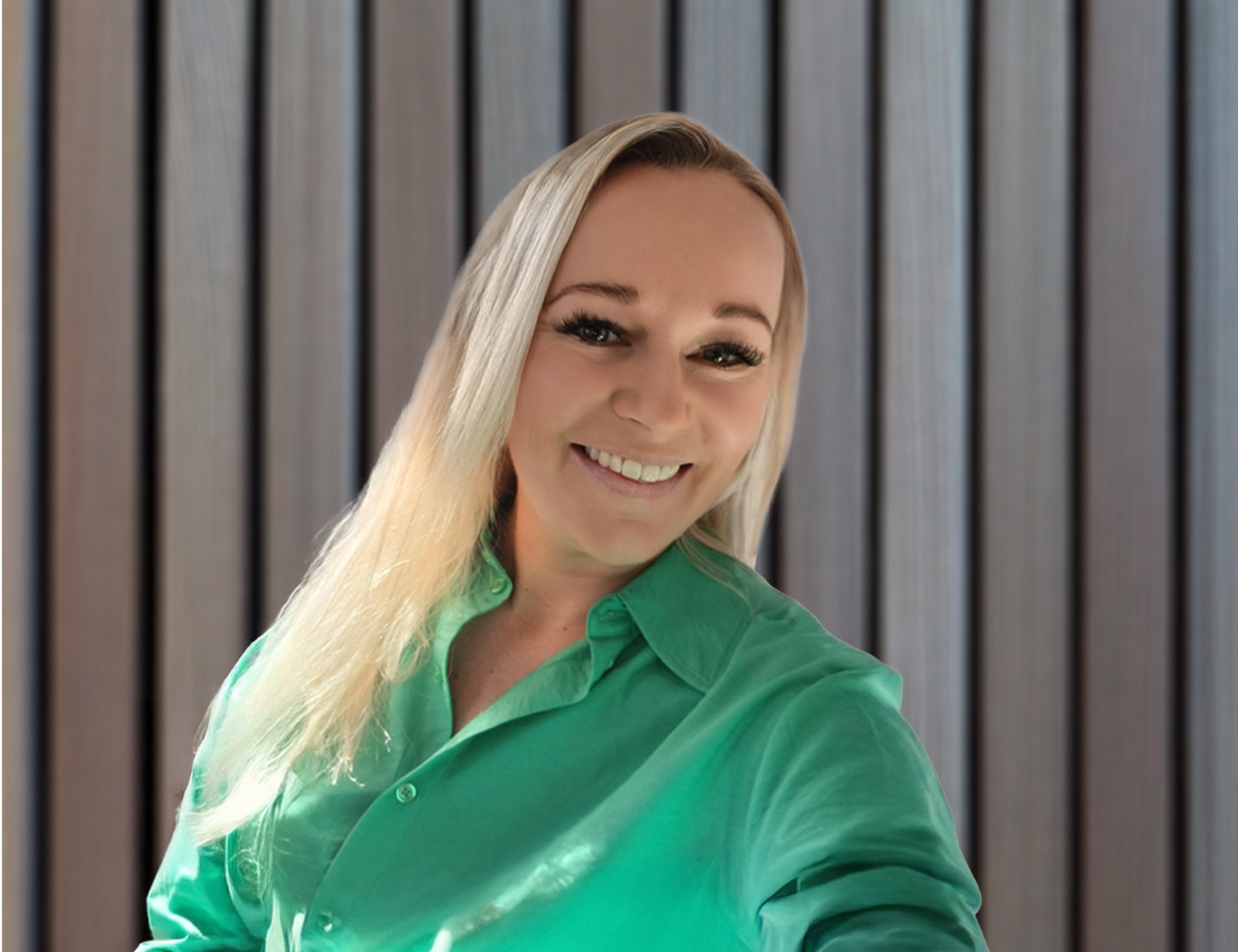 Person med långt blont hår som bär en ljusgrön skjorta, stående framför en bakgrund med vertikala träpaneler.
