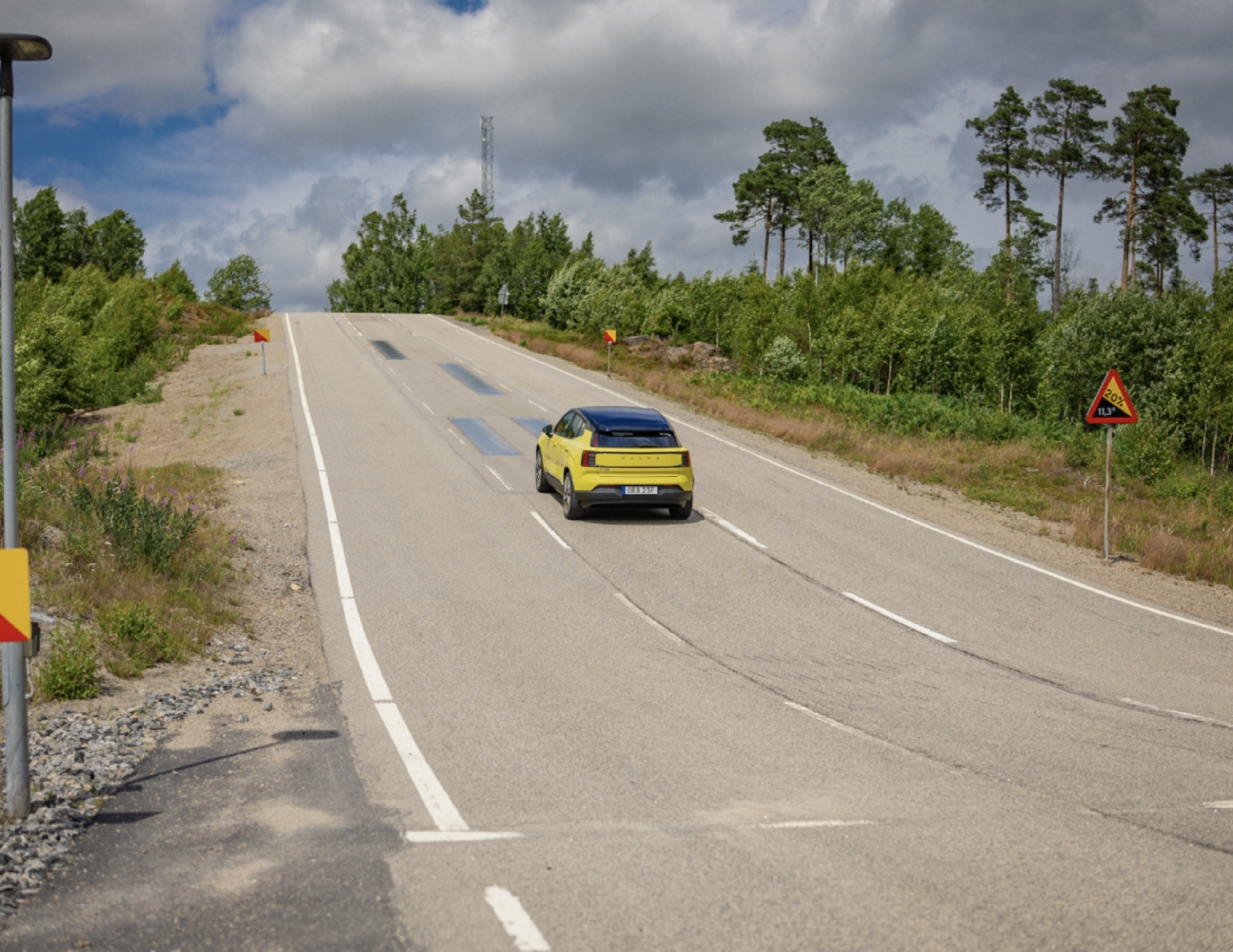 En gul Volvo C40 kör uppför en landsväg omgiven av grönska och träd under en delvis molnig himmel, med vägskyltar synliga längs vägkanten.