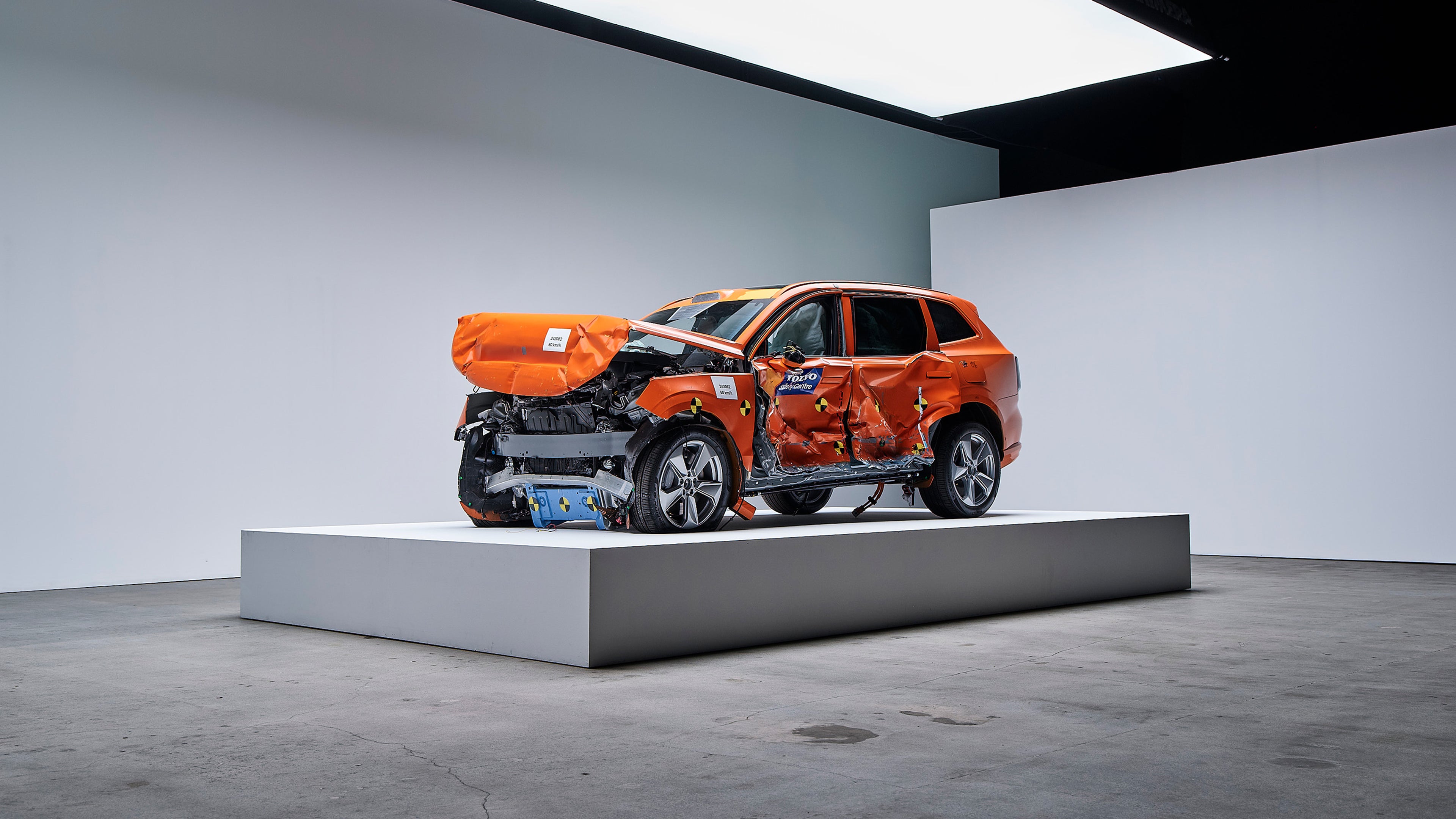 Volvo EX90 arancio sottoposta a crash test con ingenti danni anteriori e laterali esposta su una piattaforma rialzata in uno spazio espositivo minimale.