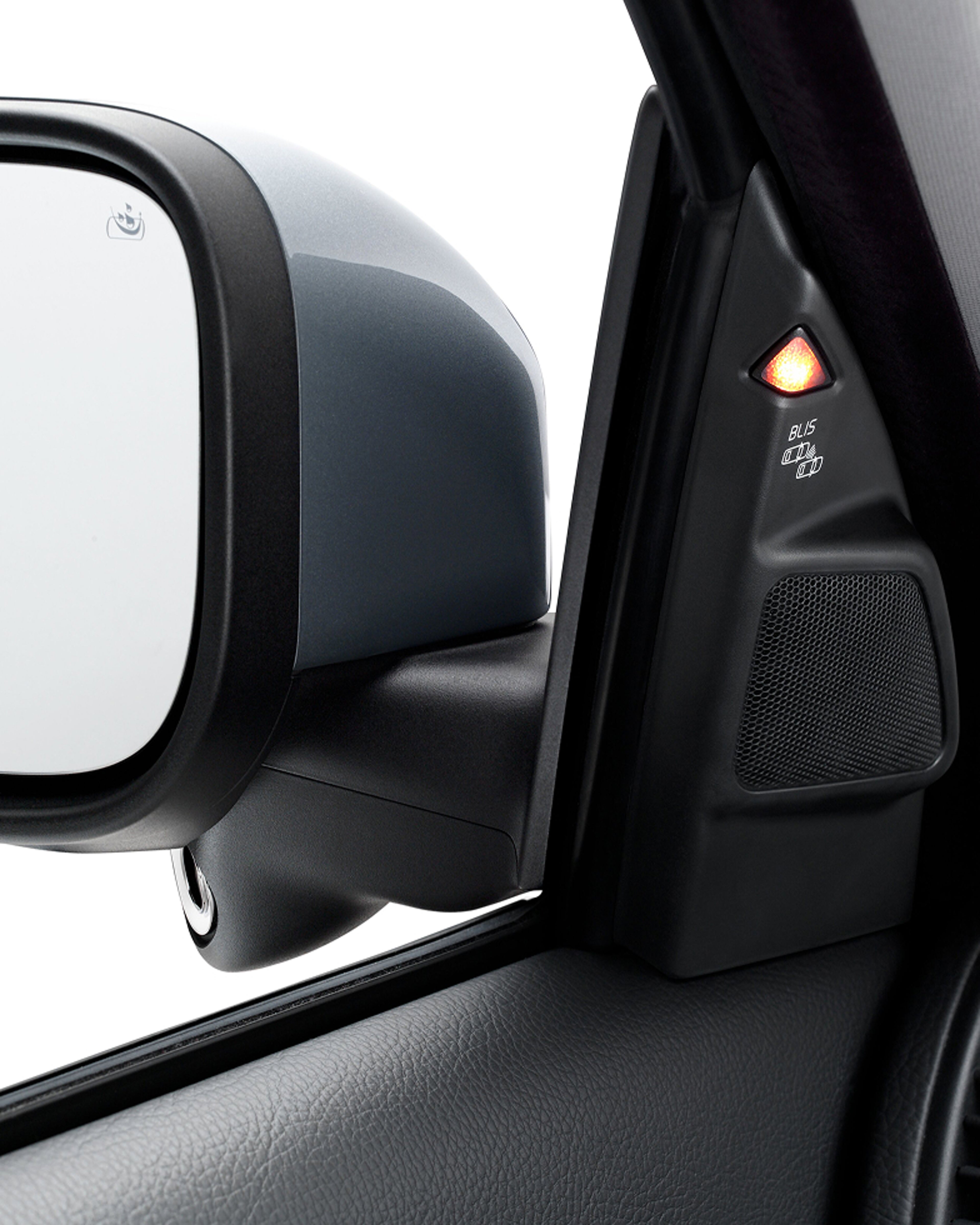 Close-up of a car’s side mirror and door pillar with an illuminated orange BLIS (Blind Spot Information System) warning light.