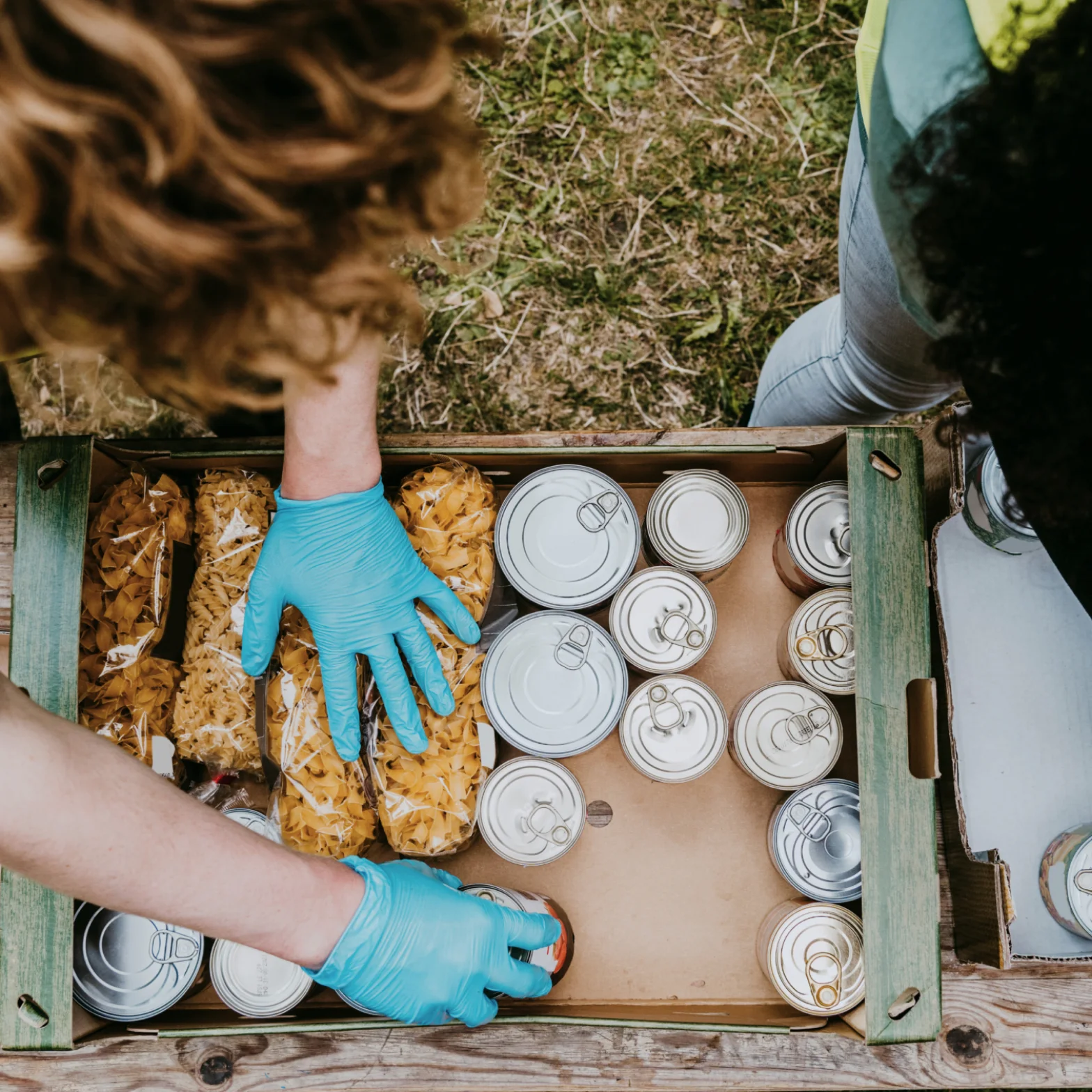 A top view of a person putting dried pasta and food tins in a box for emergency aid.
