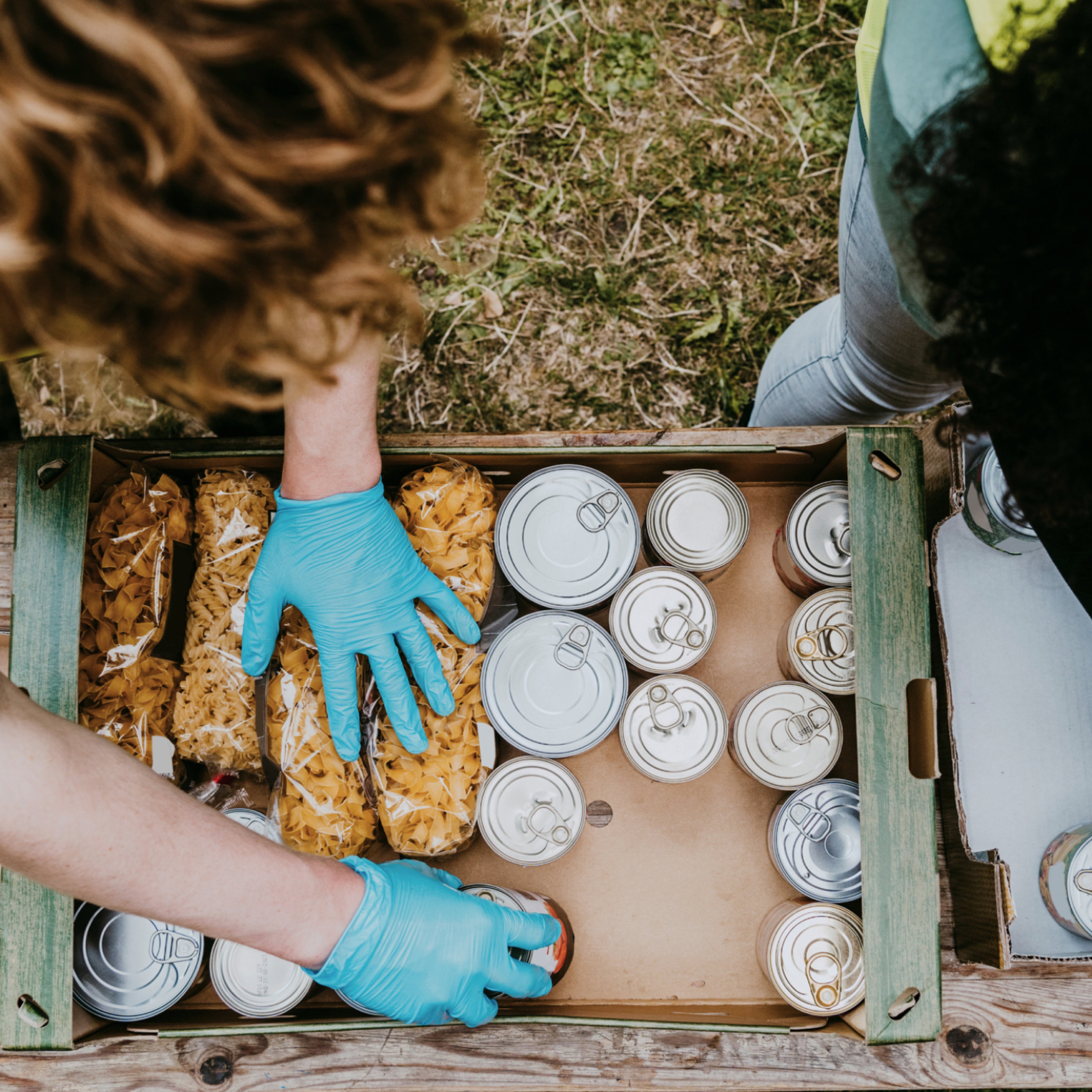 A top view of a person putting dried pasta and food tins in a box for emergency aid.
