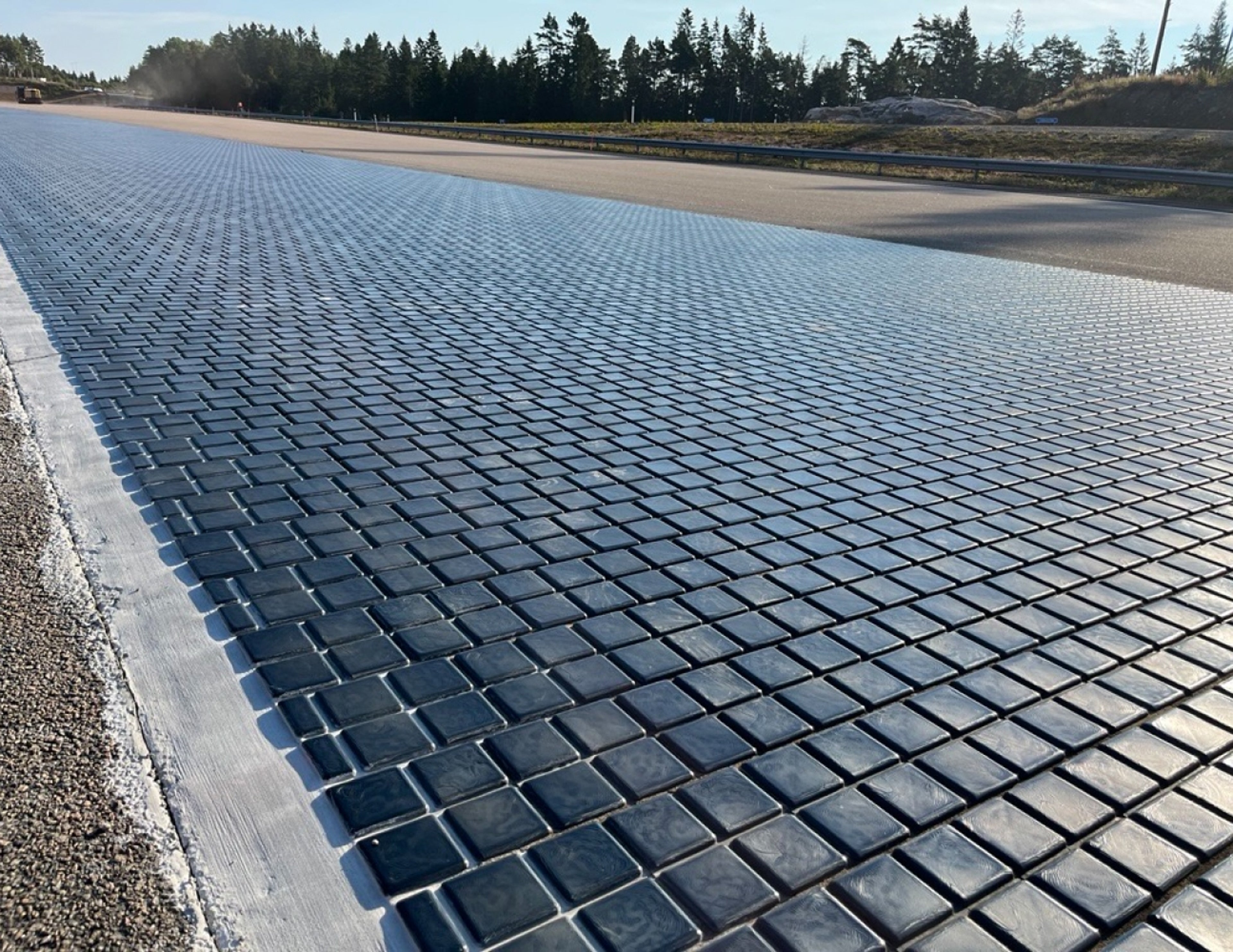 Närbild av en vägyta täckt med fyrkantiga solpaneler, som sträcker sig i fjärran med träd och en klar himmel i bakgrunden.