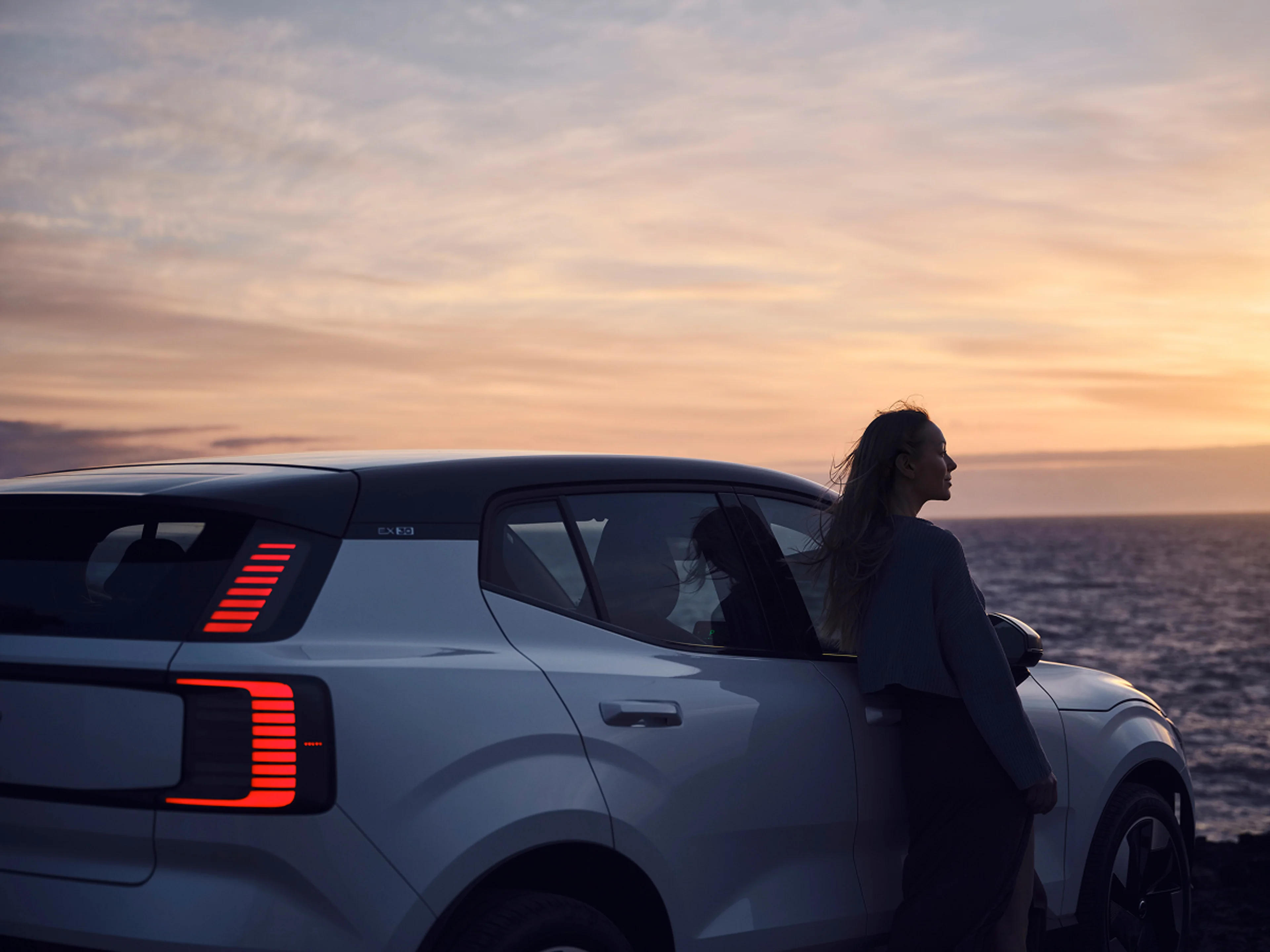 A woman stands next to a Volvo electric car parked by the ocean at sunset, symbolizing Volvo’s commitment to electrification and sustainable mobility.