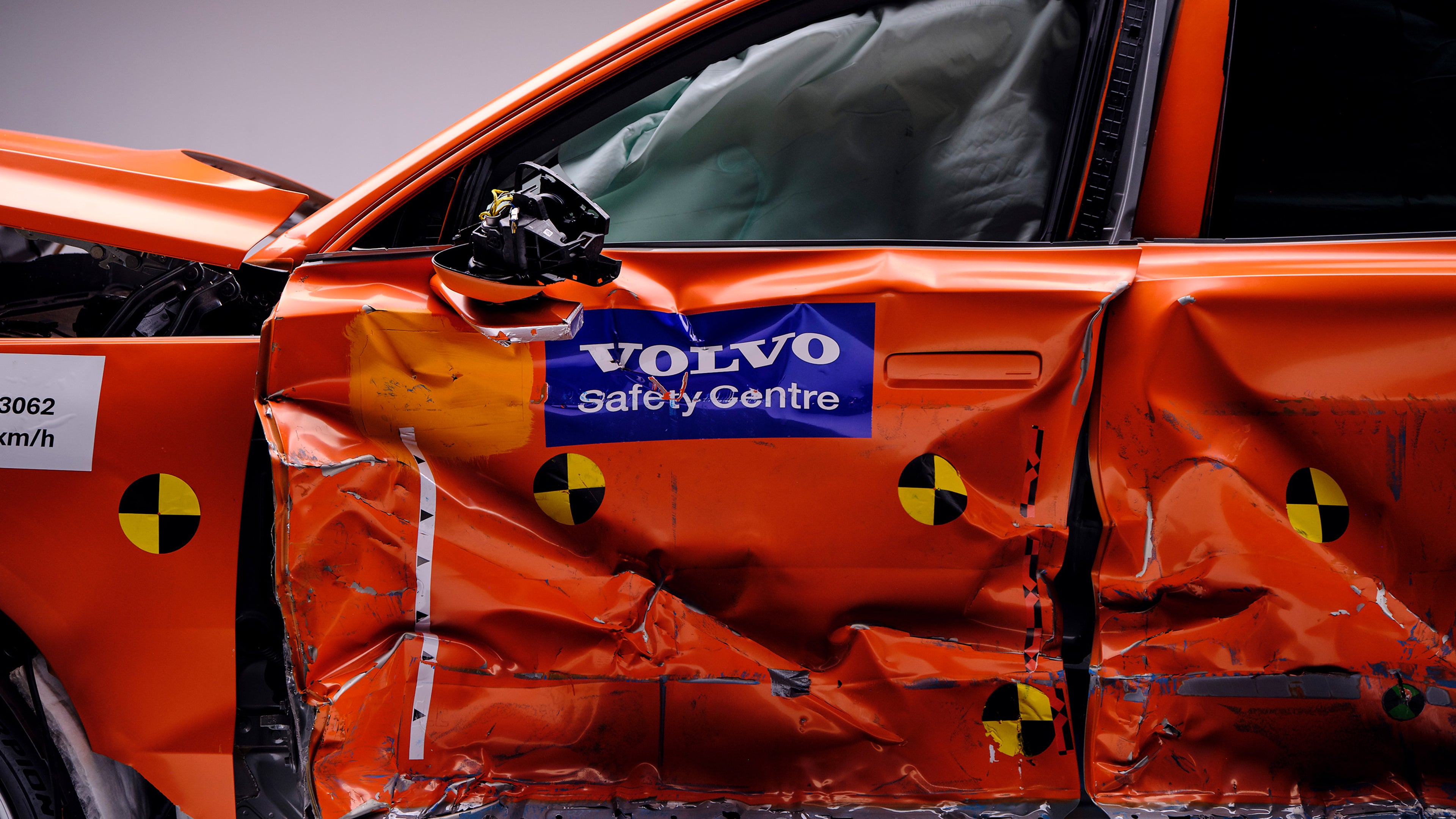 Vista laterale di un’auto Volvo arancione sottoposta a crash test con gravi danni alle portiere, indicatori per crash test ed etichetta del Safety Centre.