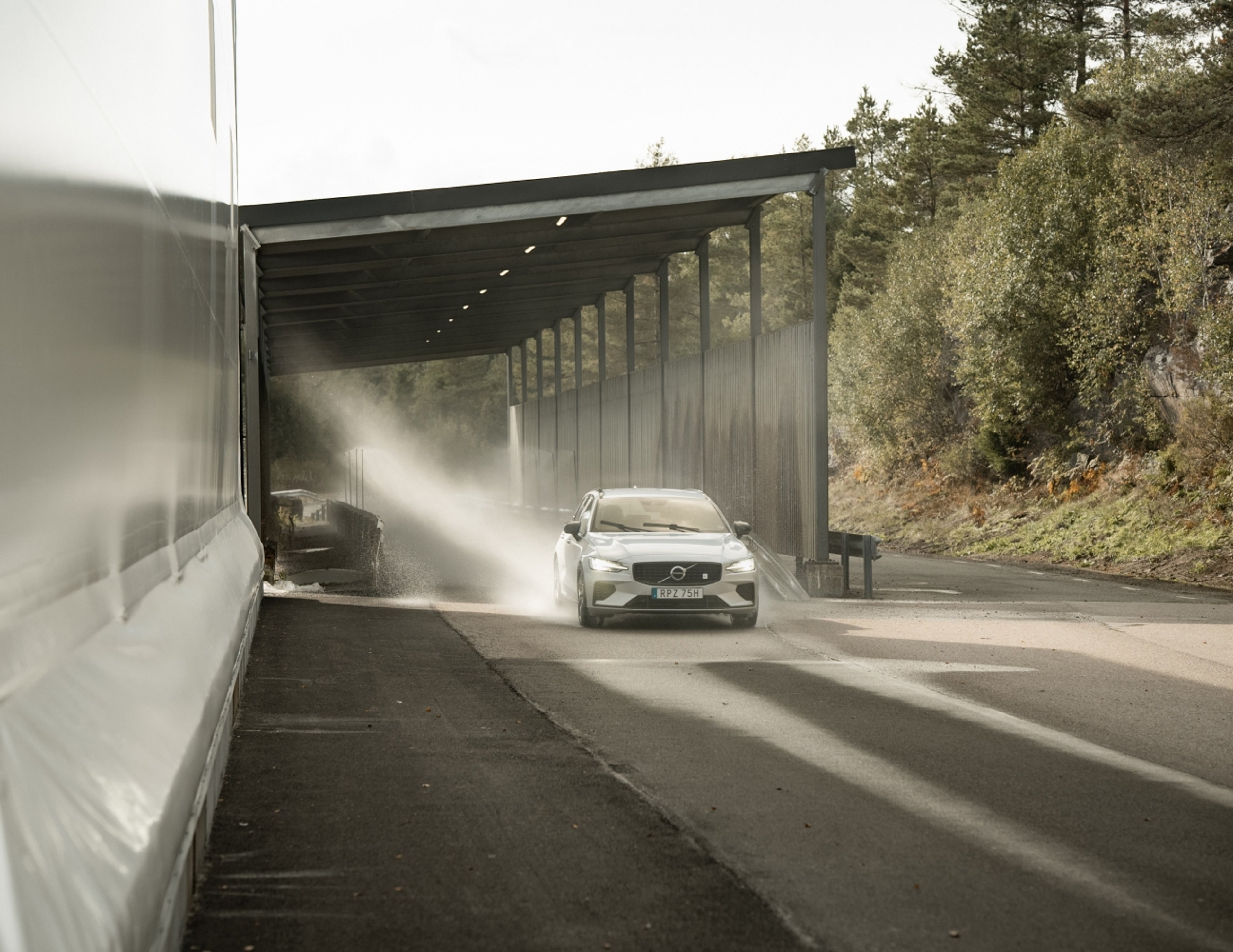 En vit Volvo V60 kör genom en vattenspraytestanläggning på en utomhusbana, med vattenstrålar som sprutar över fordonet under en täckt struktur omgiven av träd.