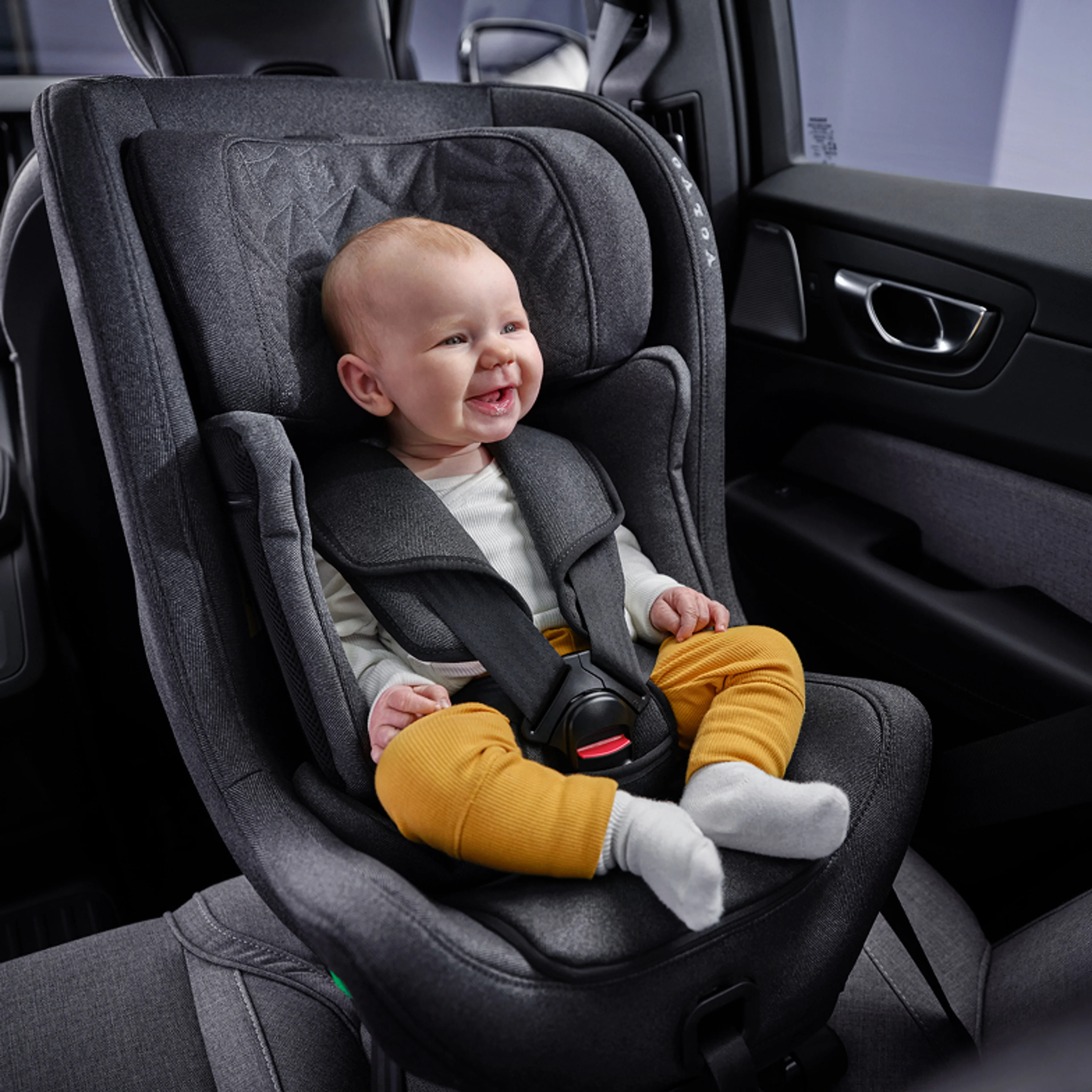 Leende bebis i vit tröja, gula leggings och vita strumpor, sittandes i en bakåtvänd bilbarnstol i en Volvo.