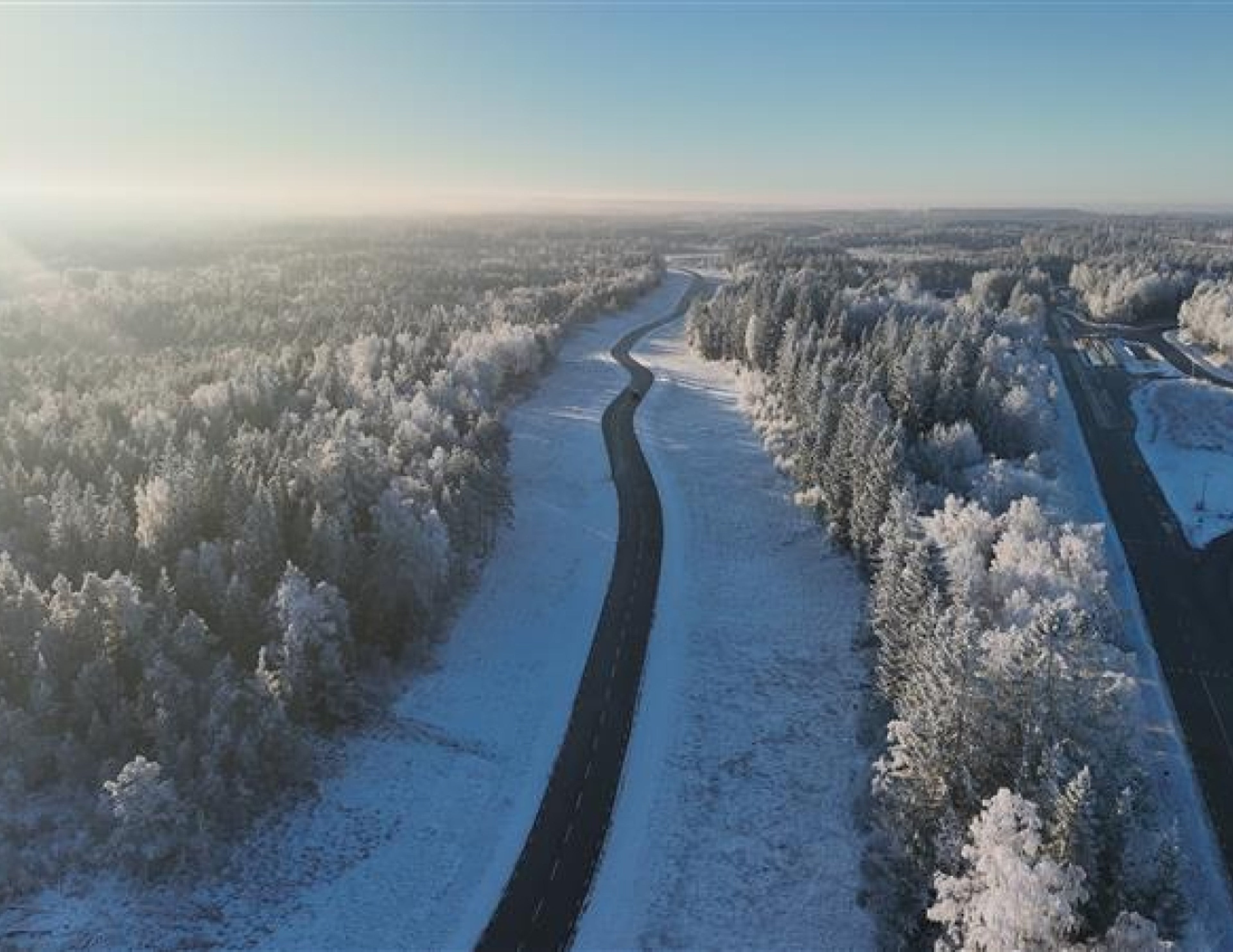 En slingrande väg skär genom ett snöigt skogslandskap under en klarblå himmel. Frosttäckta träd kantar båda sidor av vägen;