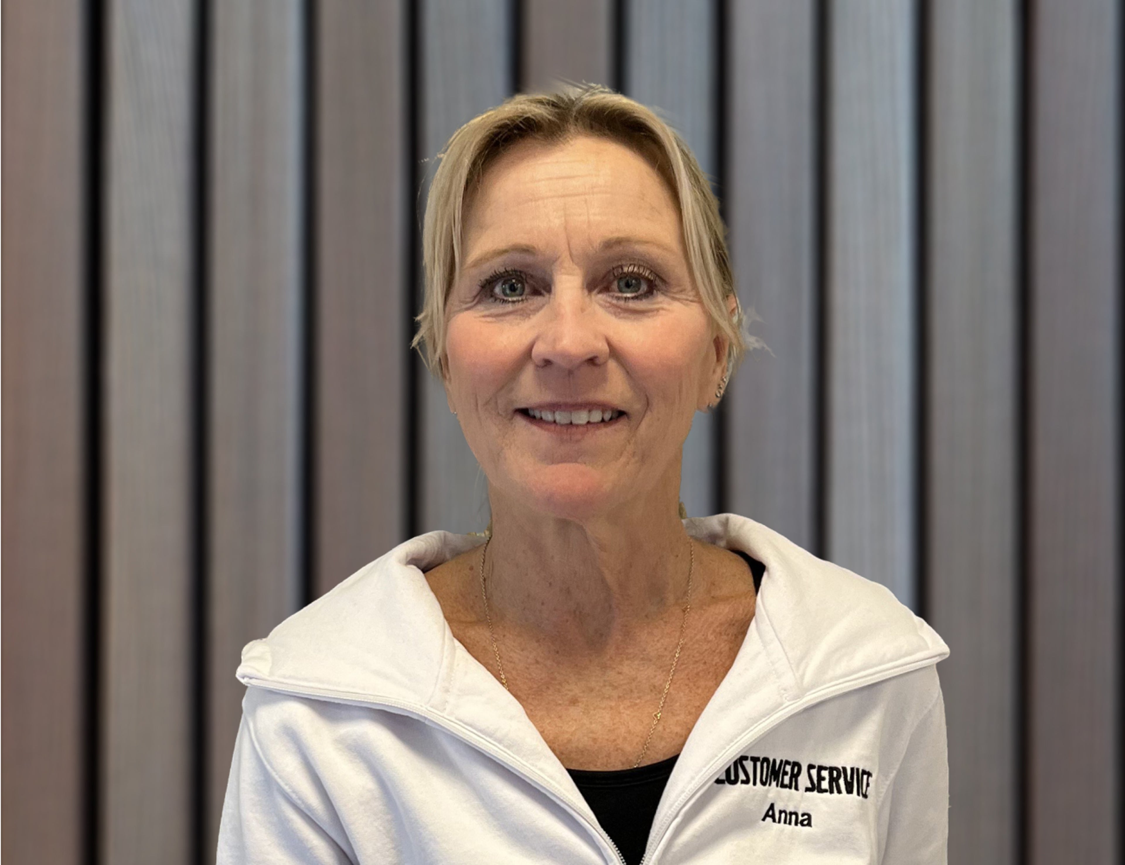 En person som bär en vit jacka med dragkedja och texten "Kundtjänst Anna" broderad på står framför en bakgrund med vertikala grå paneler.
