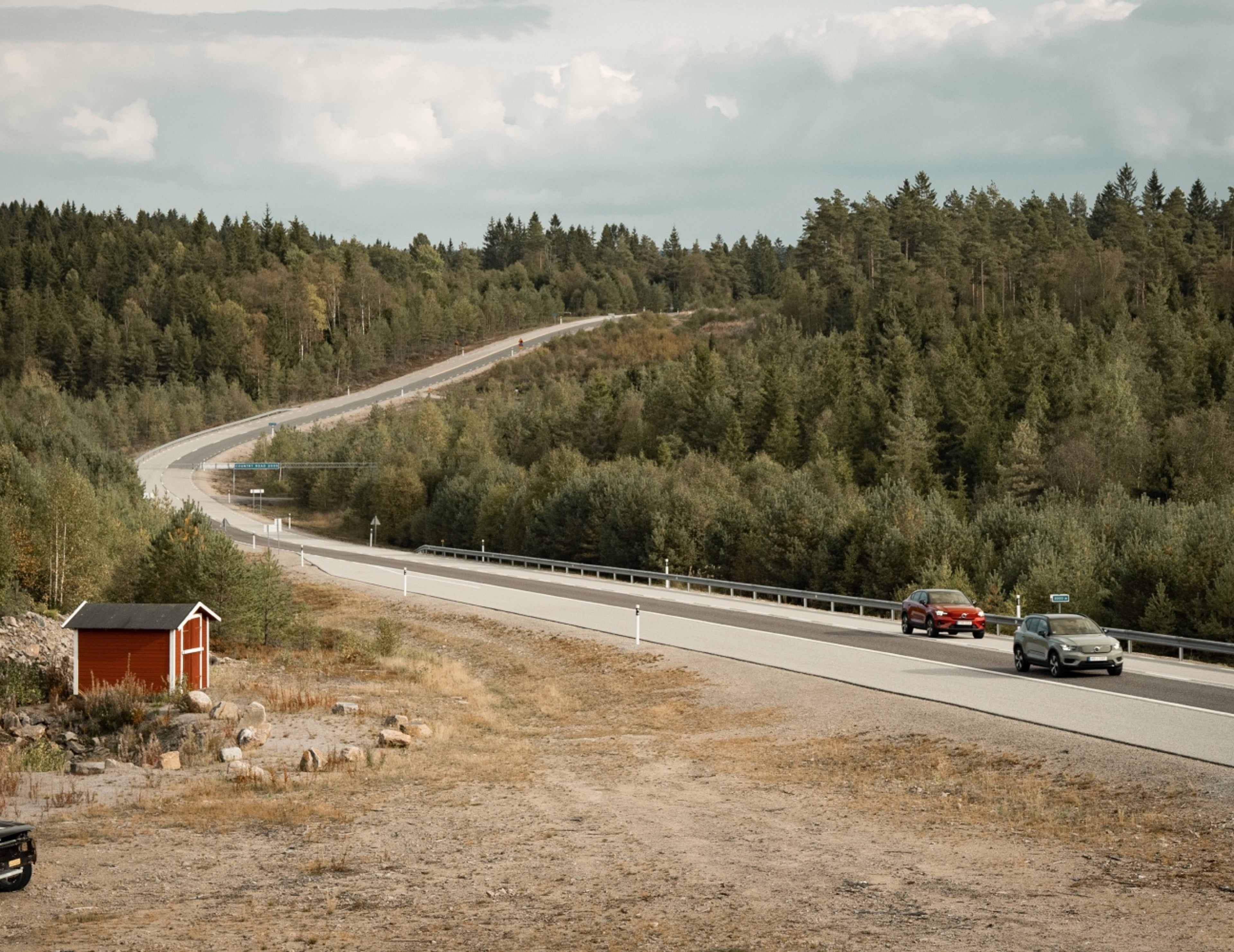 Två bilar, varav en röd Volvo S60 sedan, kör längs en slingrig landsväg kantad av tät skog och ett litet rött skjul under en molnig himmel.