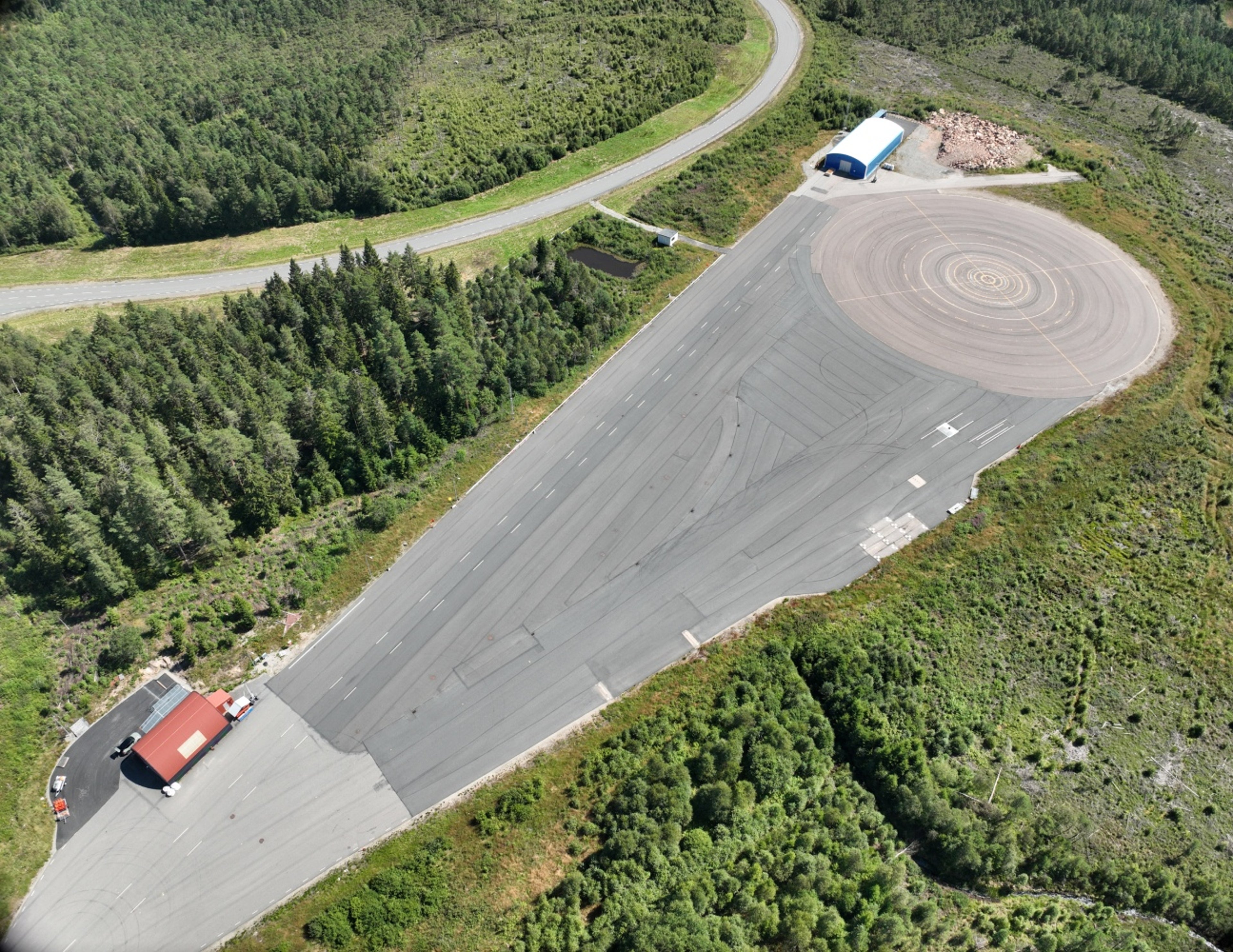 Flygfoto över en stor testbana för fordon omgiven av grönska, med en cirkulär halkbana, raka körfält och två små byggnader i motsatta ändar av anläggningen.