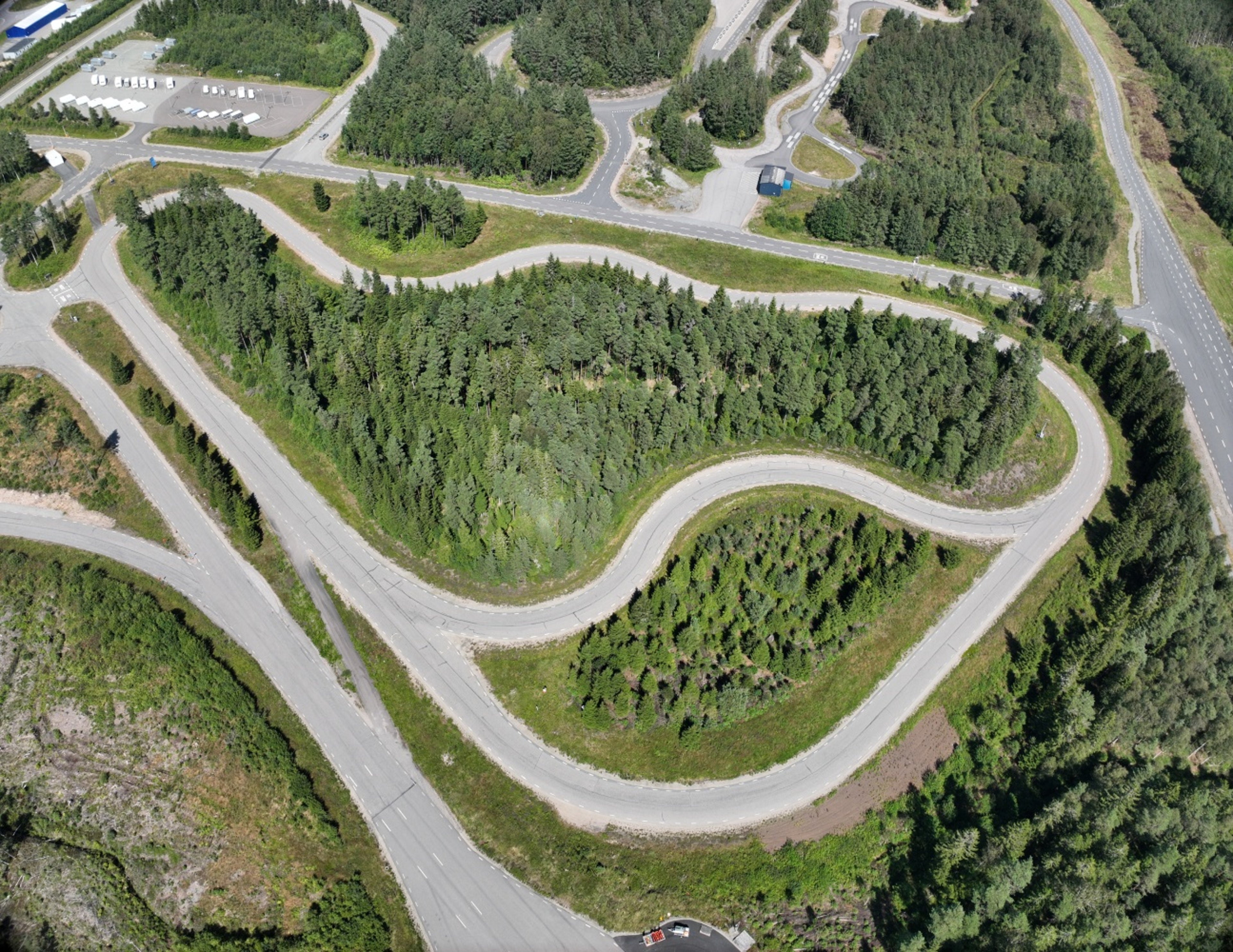 Flygfoto över en slingrande testbana omgiven av tät grön skog och flera vägar, utan synliga fordon på banan eller närliggande vägar.