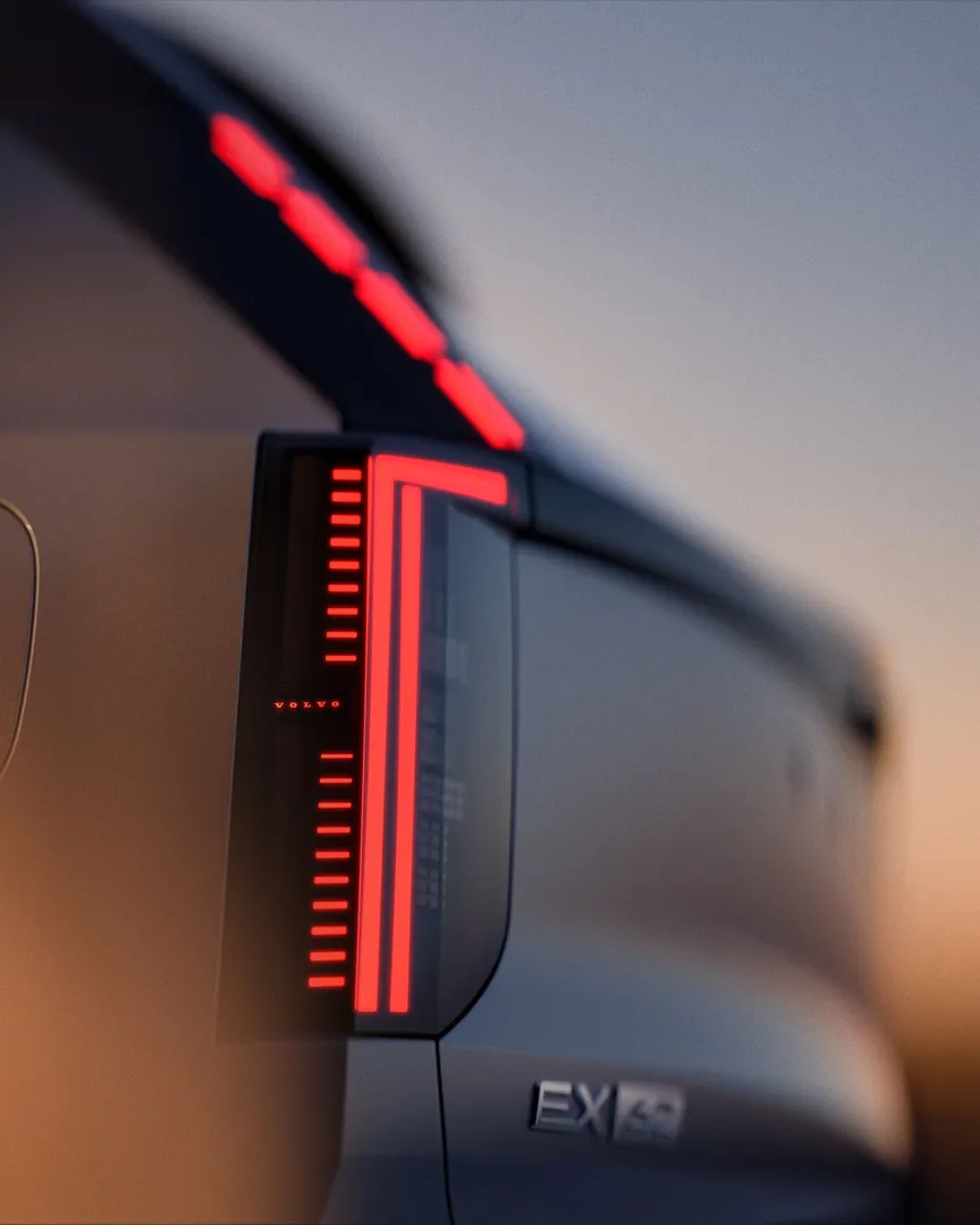 The rear left corner of the fully electric Volvo EX60. The taillight glows in red and orange tones, while the rest of the car appears as a silhouette against a warm orange background.