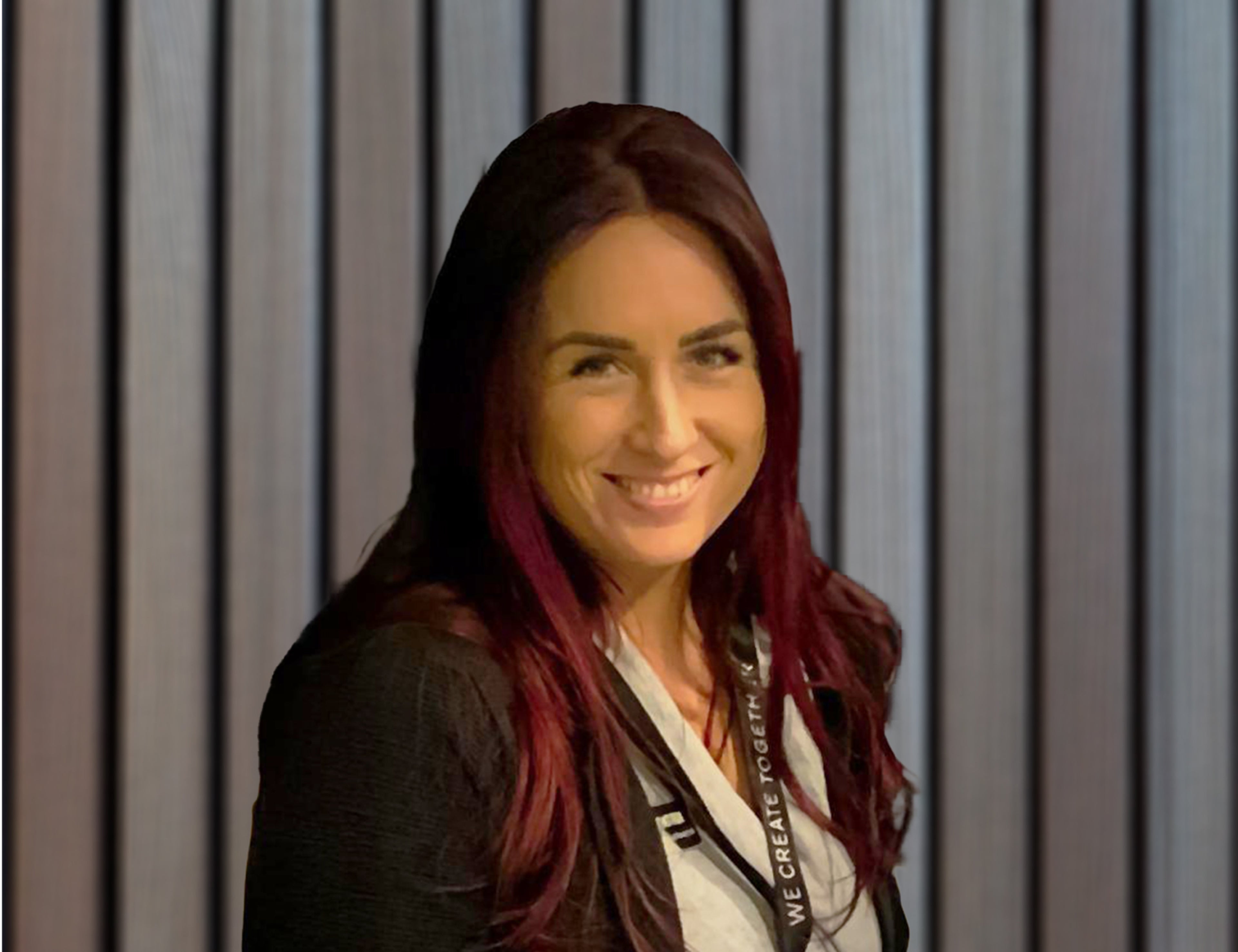 En person med långt, mörkrött hår står framför en vertikal grå panelvägg, klädd i en mörk kavaj och ett nyckelband med vit text.