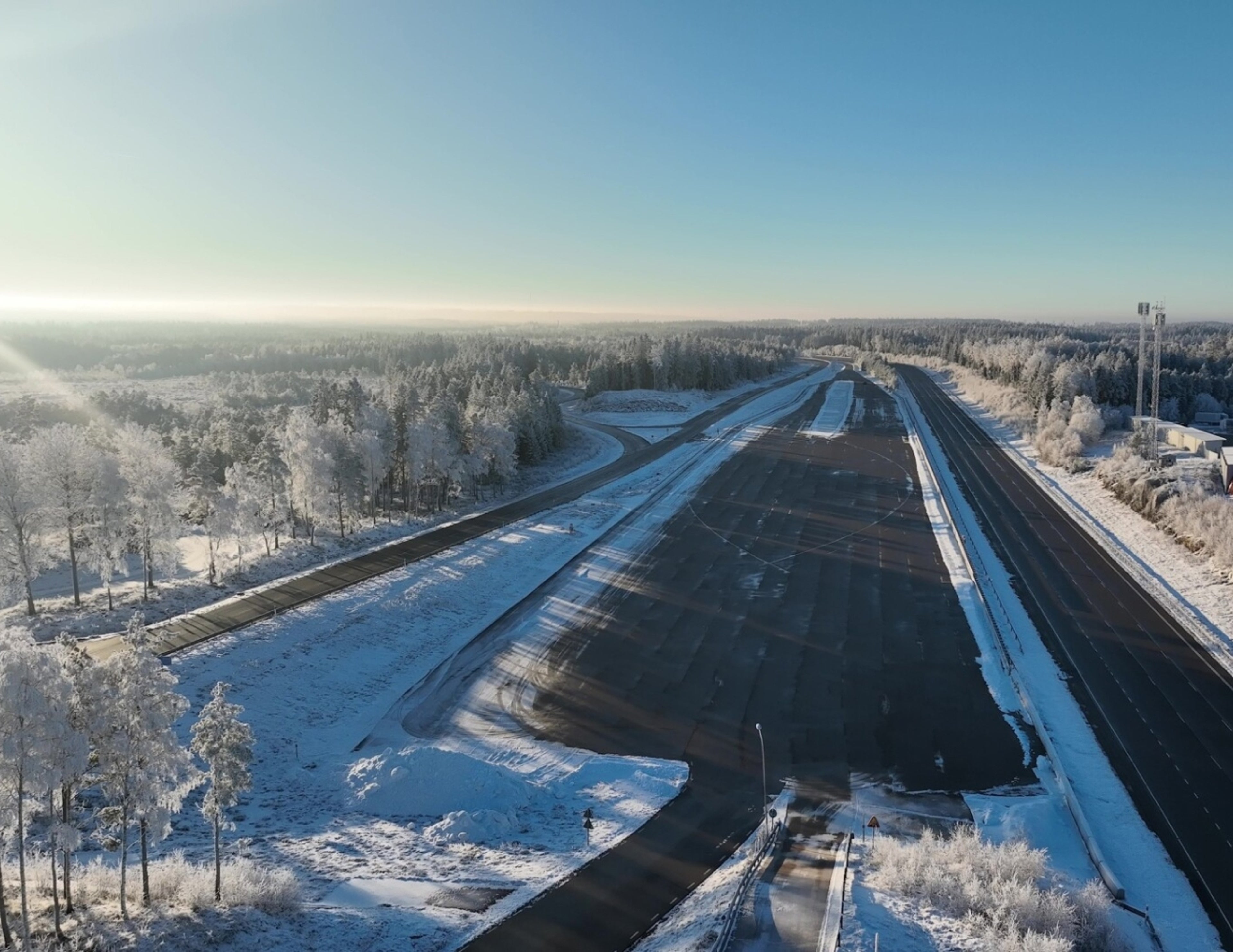 Flygfoto över en snötäckt testbana omgiven av frostiga träd och klarblå himmel, med tomma asfaltsfiler och vinterlandskap som sträcker sig i fjärran.