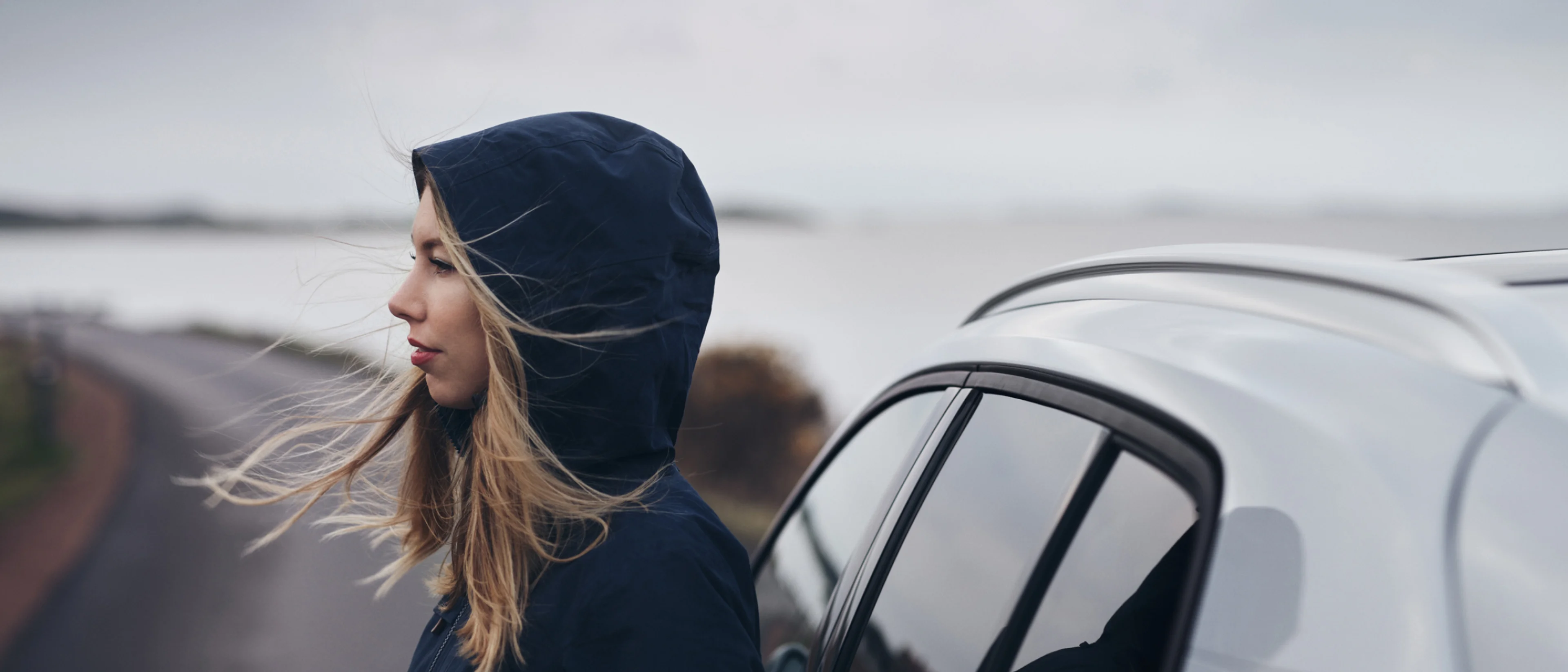 Side profile shot of a woman in a blue hoodie jacket stood beside a white Volvo.