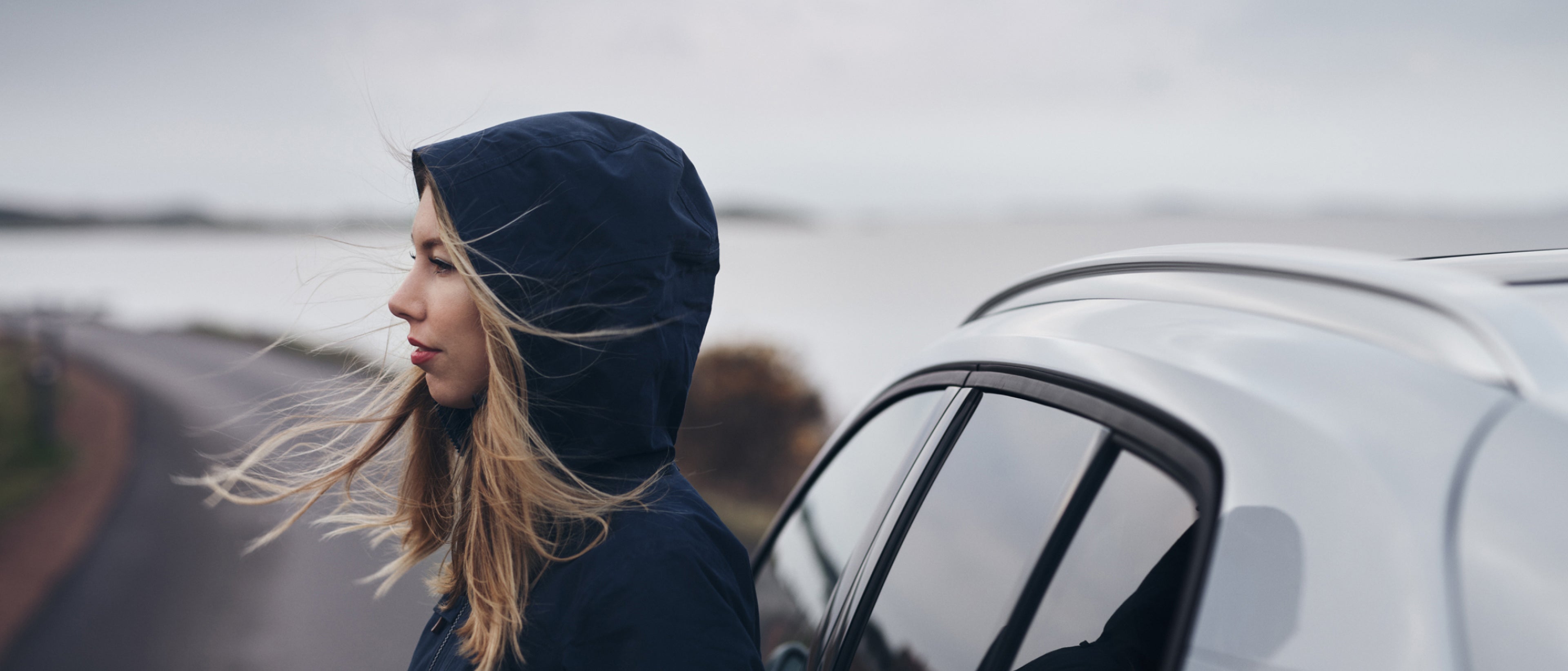 Plano de perfil lateral de una mujer con una sudadera azul con capucha de pie junto a un Volvo blanco.