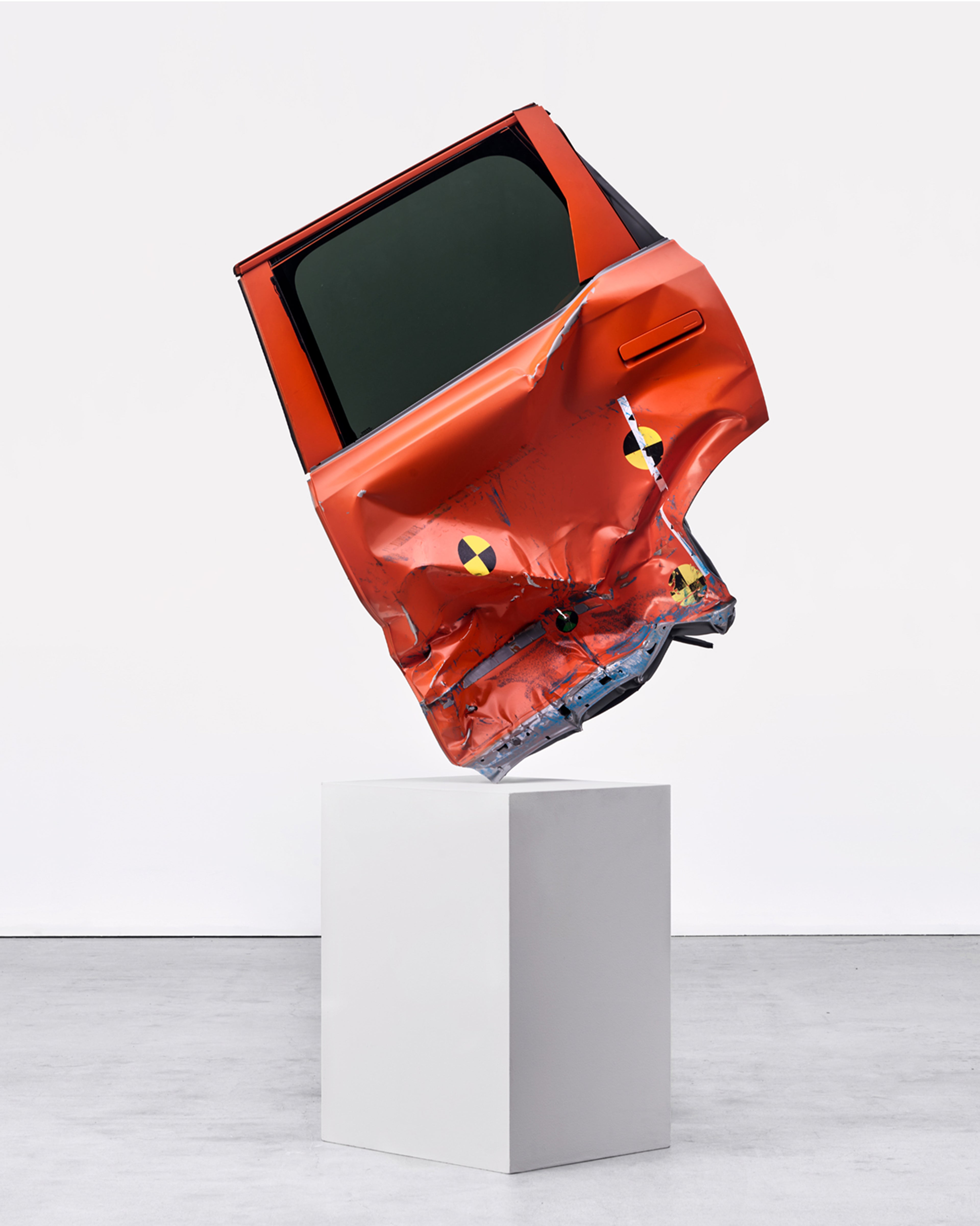 A crumpled orange car door with crash test markers displayed upright on a white pedestal in a gallery space.