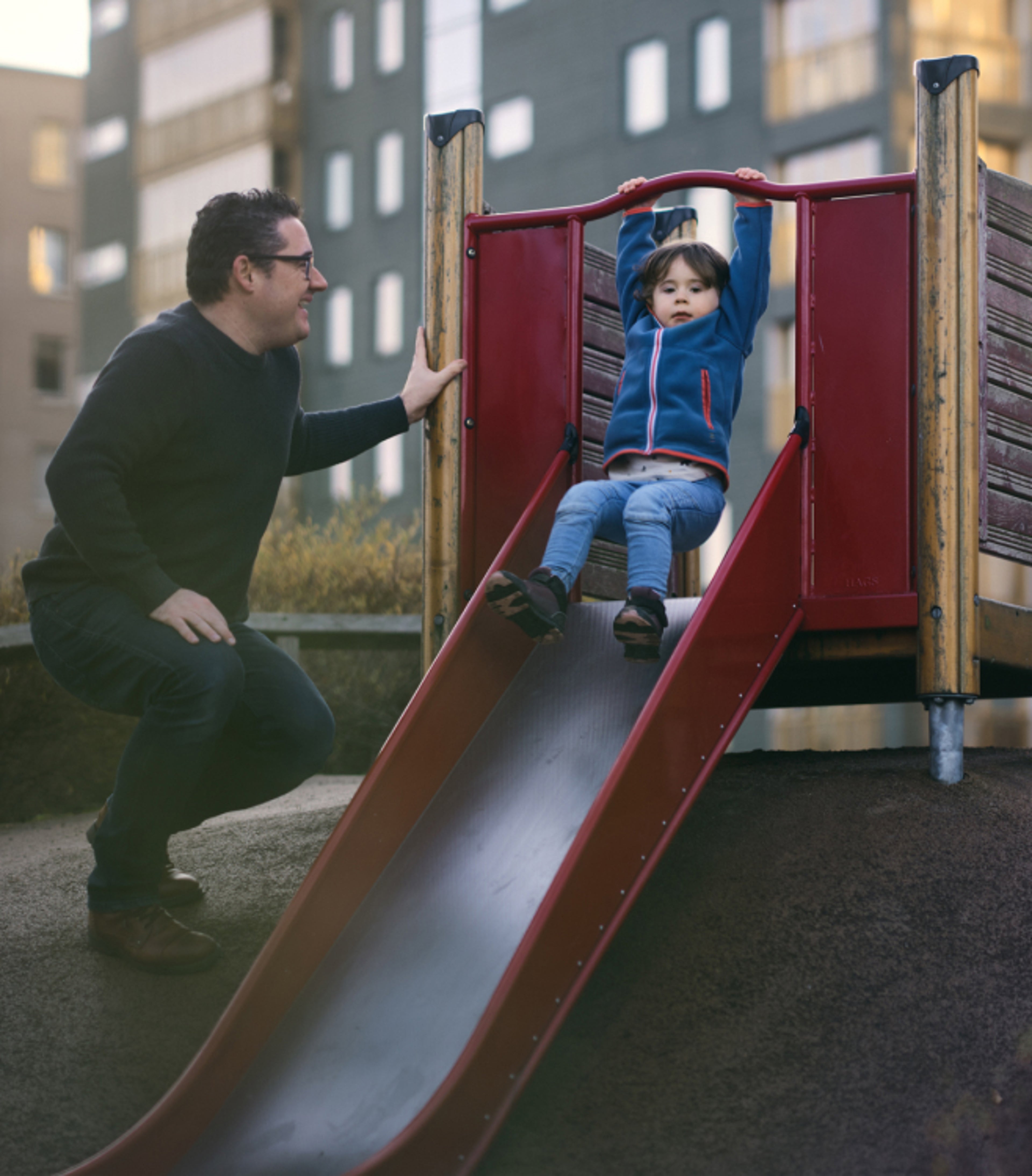 A man smiling at a toddler coming down a slide in a play area for kids.