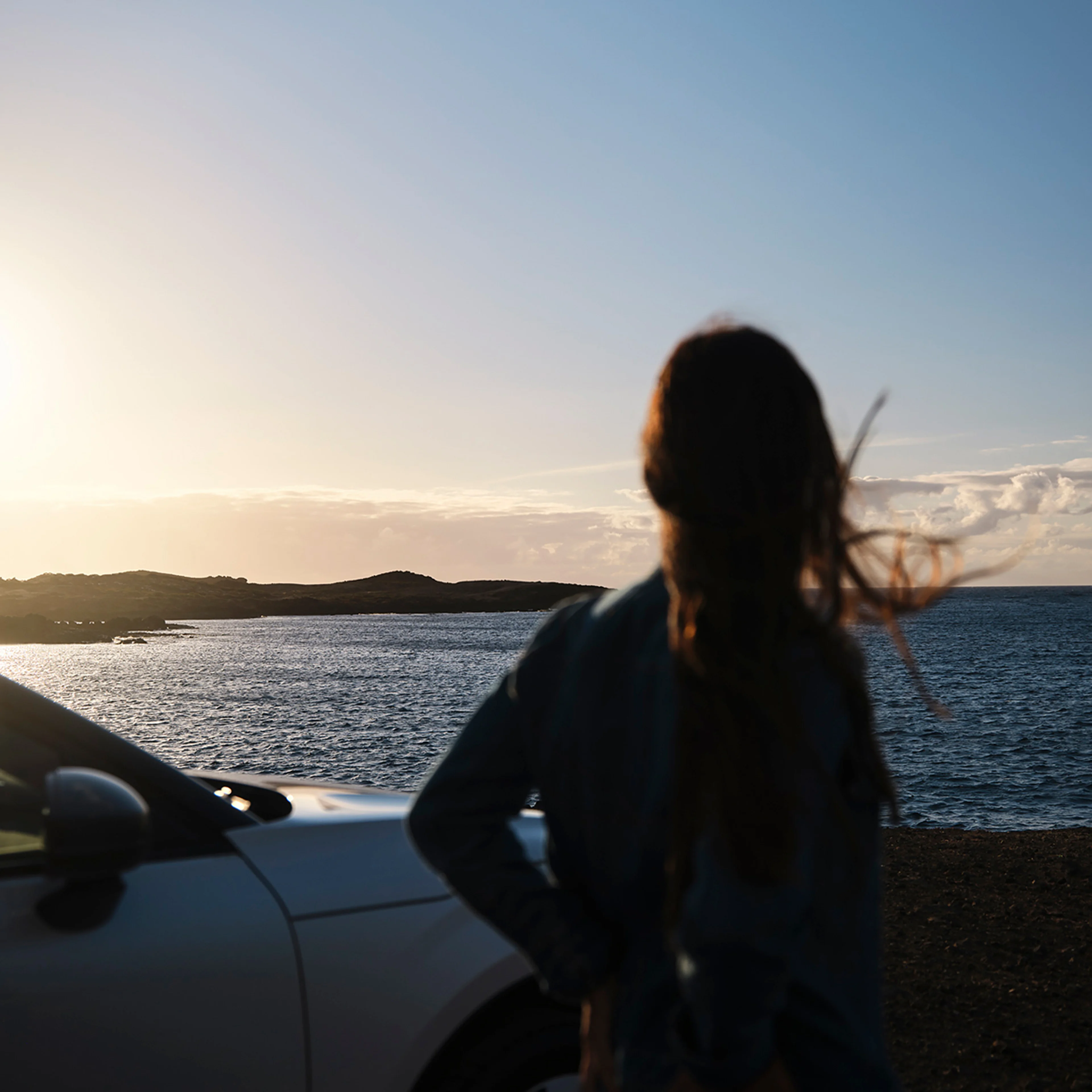 A woman stands beside a Volvo car, gazing at the ocean horizon during sunset. Symbolizing Volvo’s commitment to ocean health and its partnership with The Ocean Race.
