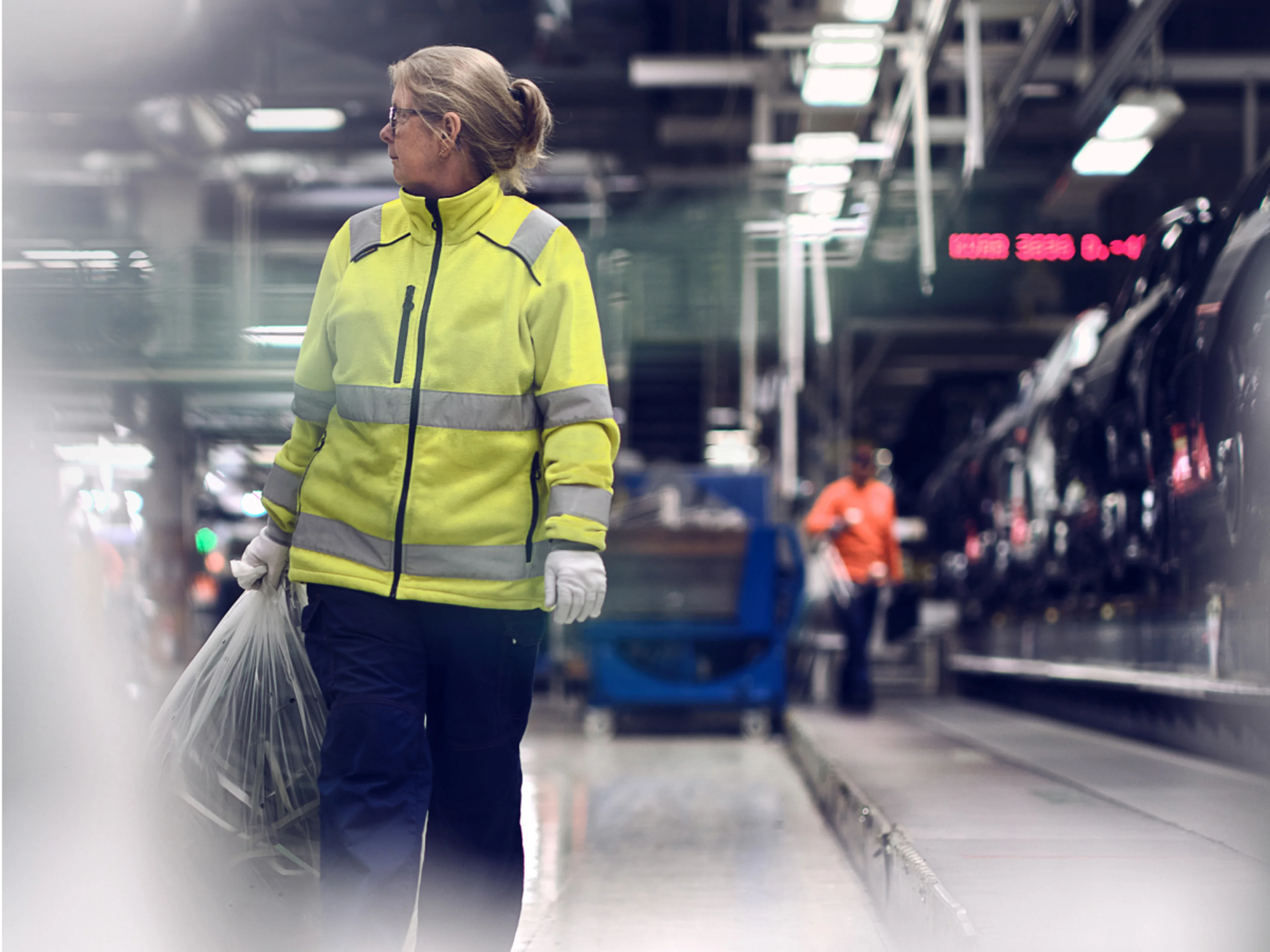 Eine Frau im Arbeitsoverall trägt eine Plastiktüte und dreht sich nach rechts um.