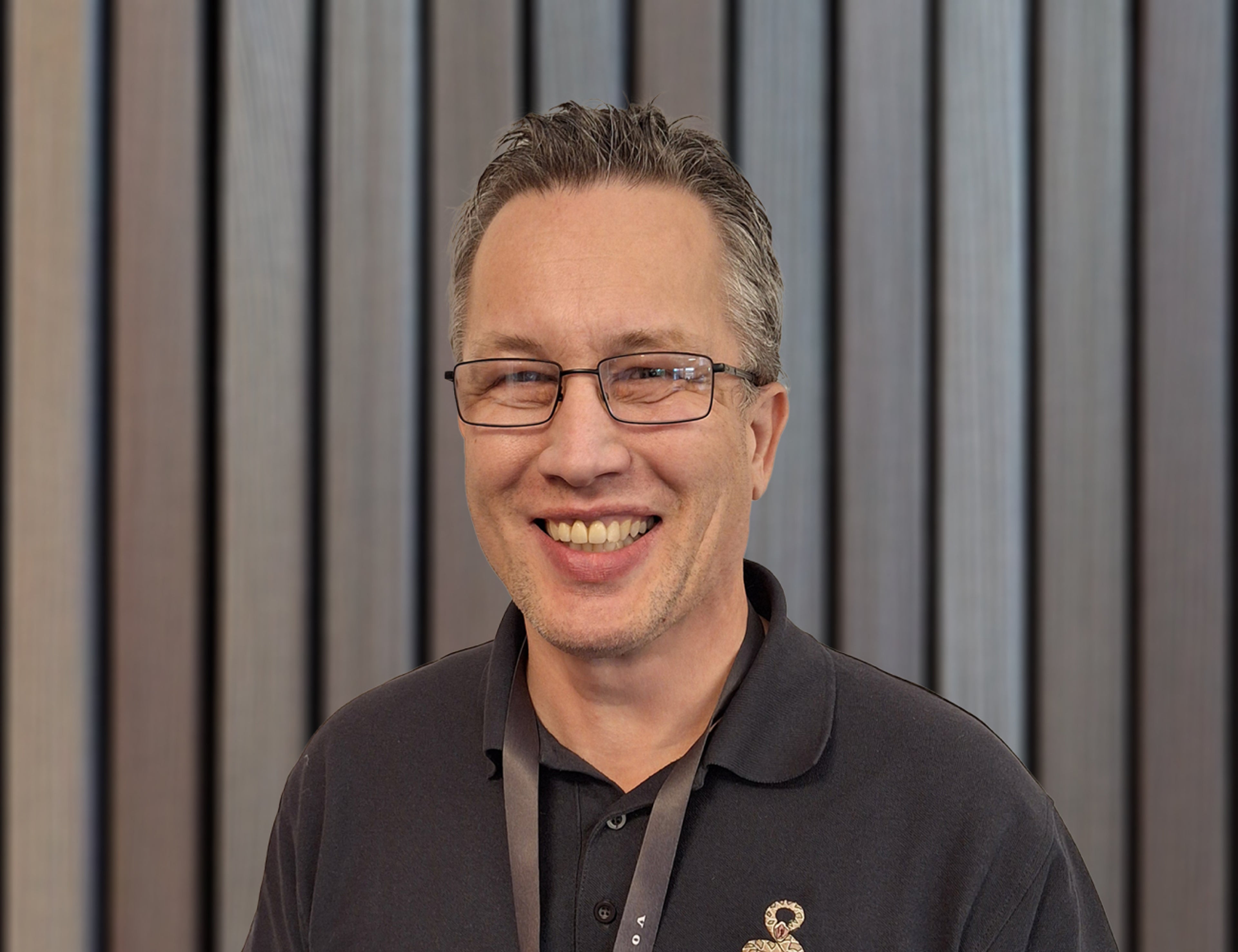 Person som bär en skjorta med svart krage med ett broderat emblem och ett nyckelband, framför en bakgrund med vertikala träpaneler.