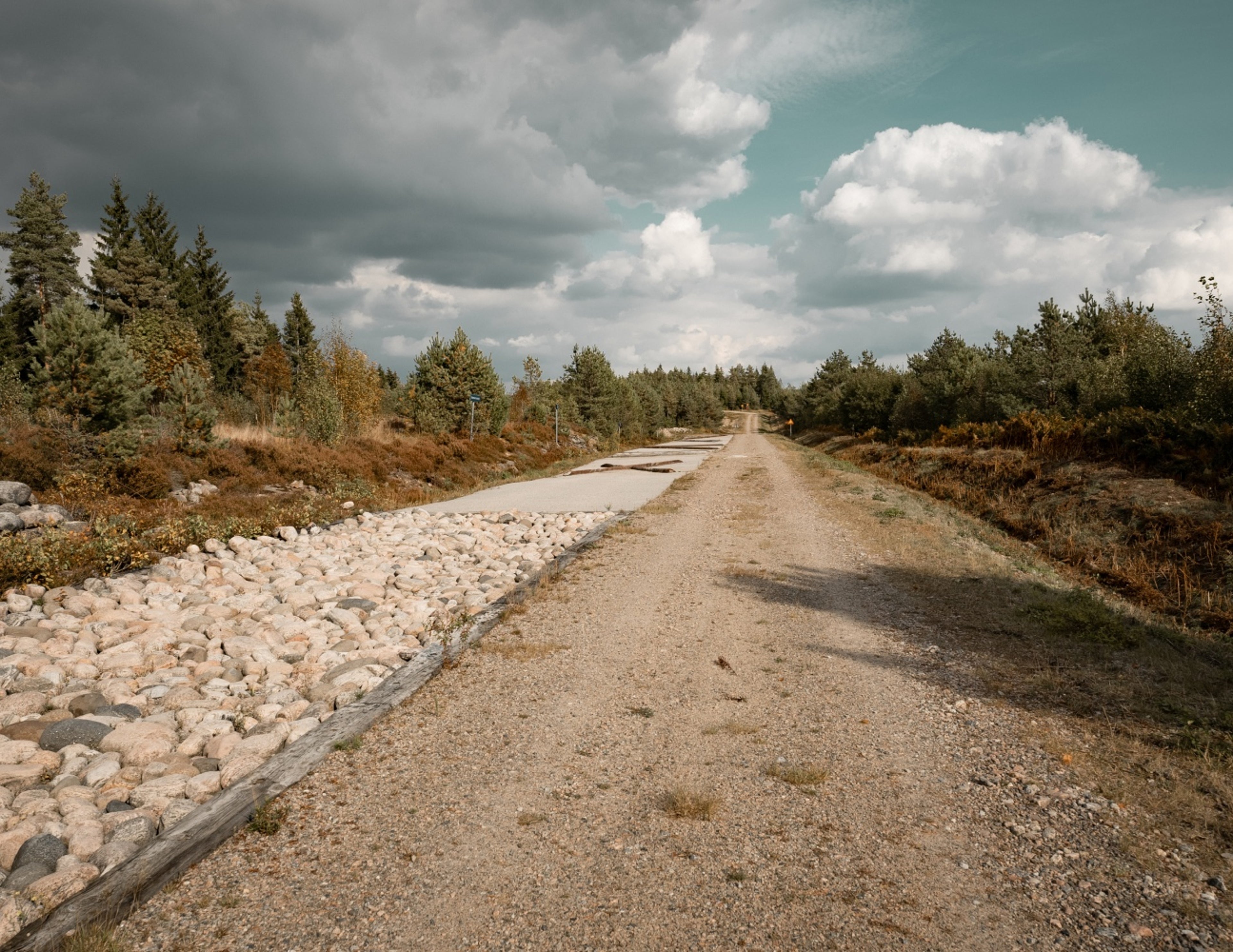 En lantlig grusväg sträcker sig i fjärran, kantad av stenar och täta gröna träd under en delvis molnig himmel.