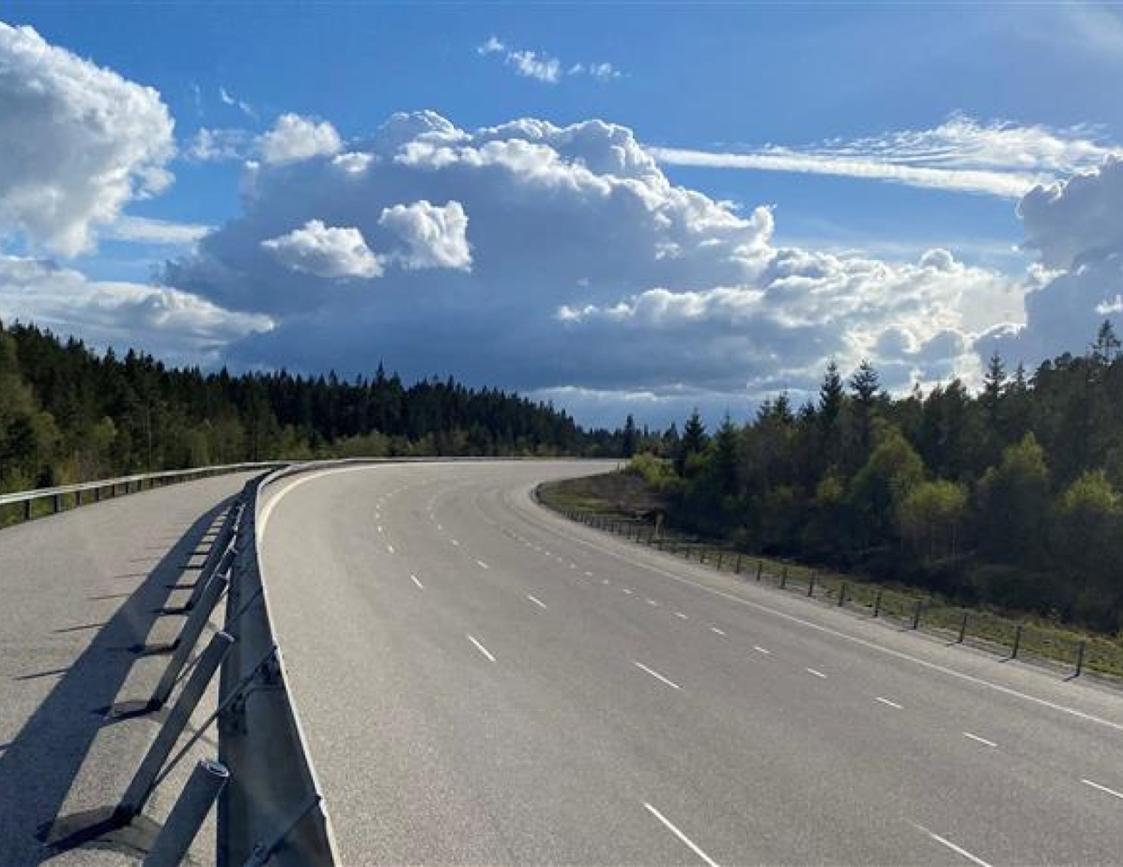 En bred, tom motorväg slingrar sig genom ett skogslandskap under en delvis molnig himmel. Inga fordon eller människor är synliga; Scenen är fridfull och öppen.