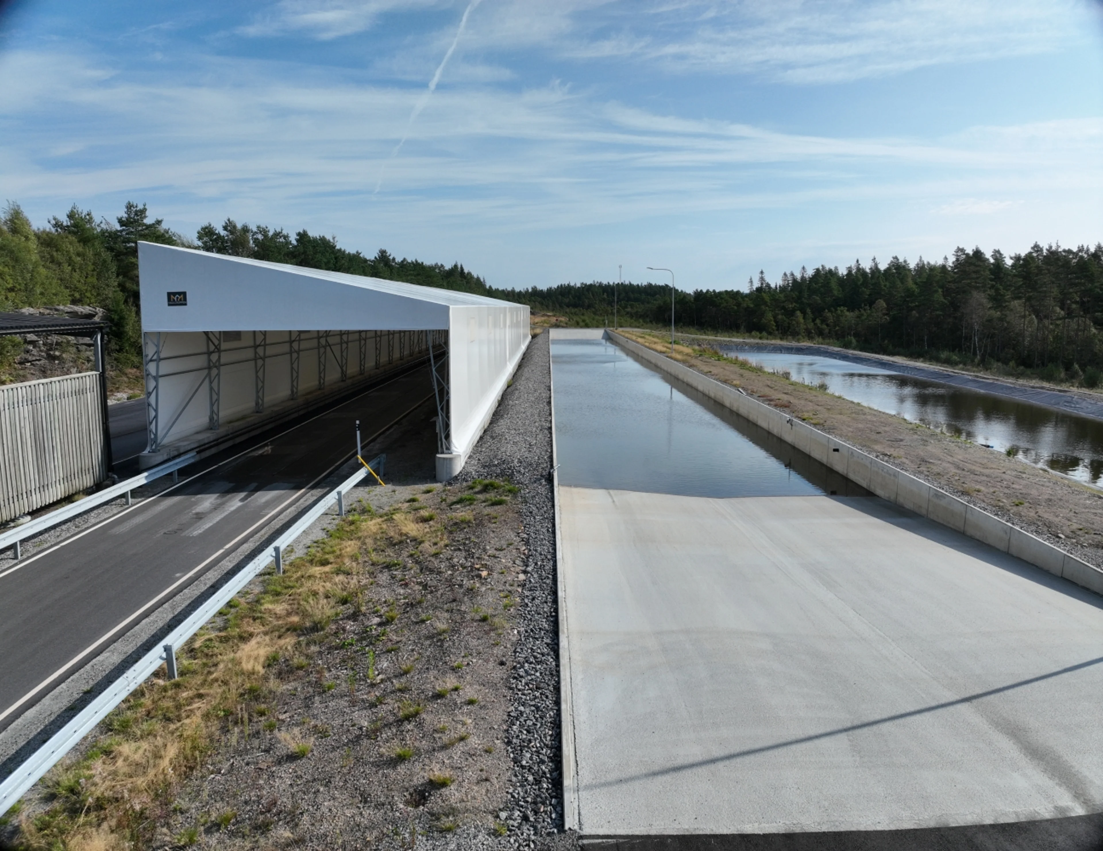 Testanläggning för bilar utomhus med en täckt testbana till vänster och en stor vattenpool till höger, omgiven av träd och bar himmel under dagsljus.