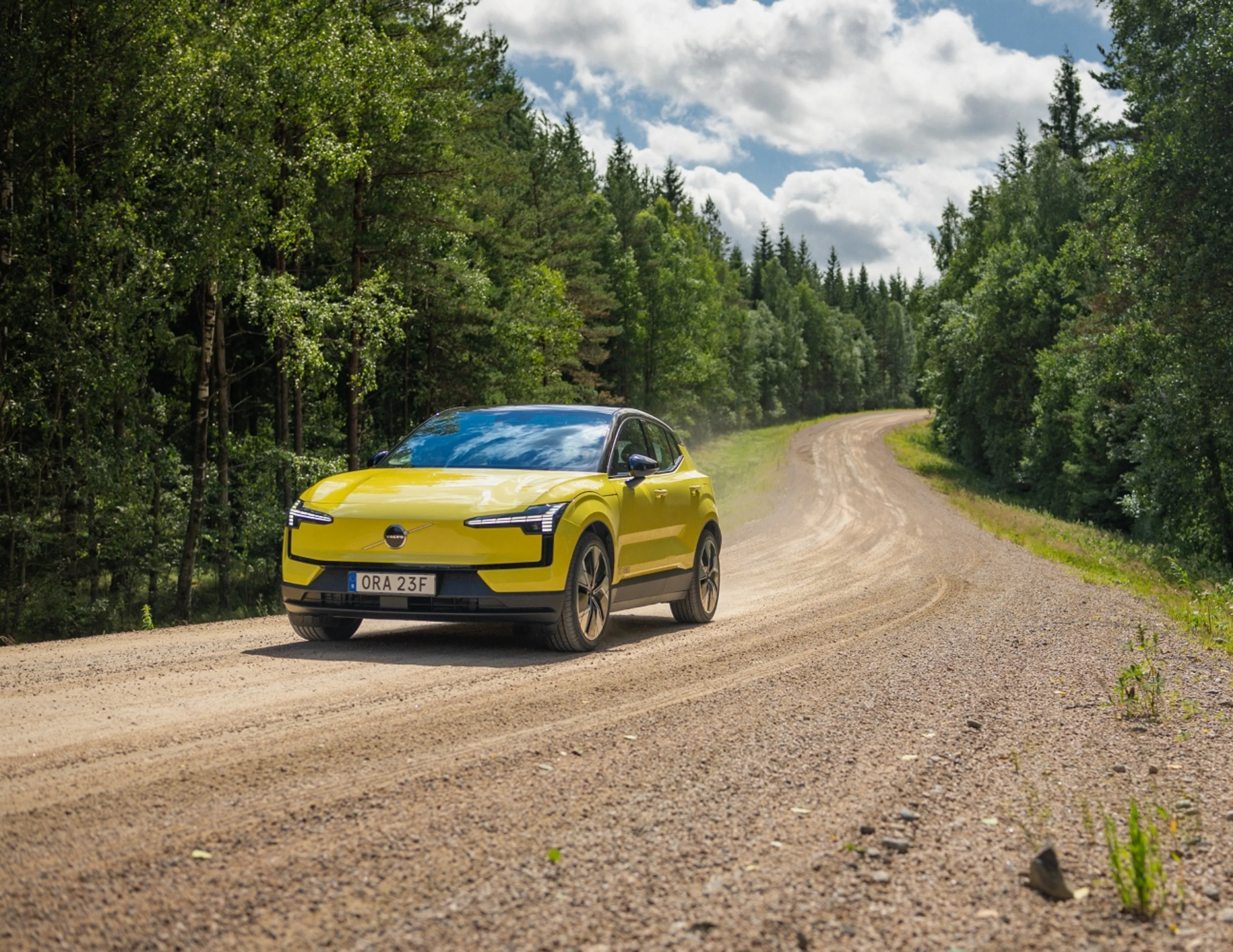 En gul Volvo EX30 kör på en slingrig grusväg genom en grönskande skog under en delvis molnig himmel och kastar upp damm när den rör sig längs den natursköna vägen.
