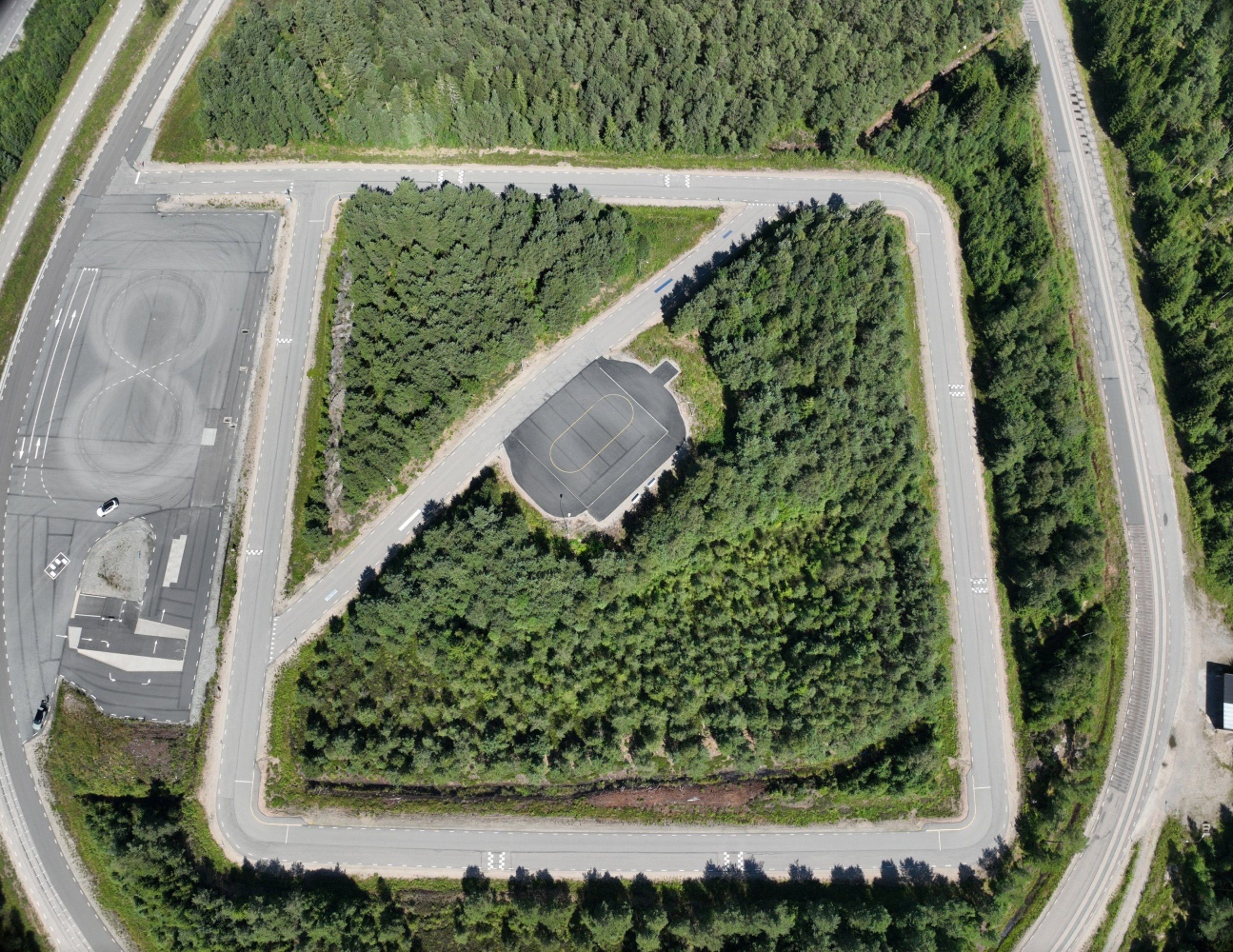 Flygfoto över en skogsbevuxen testbana med slingrande vägar som omger ett centralt asfaltsområde. Flera fordon står parkerade till vänster och täta träd fyller det inre utrymmet.