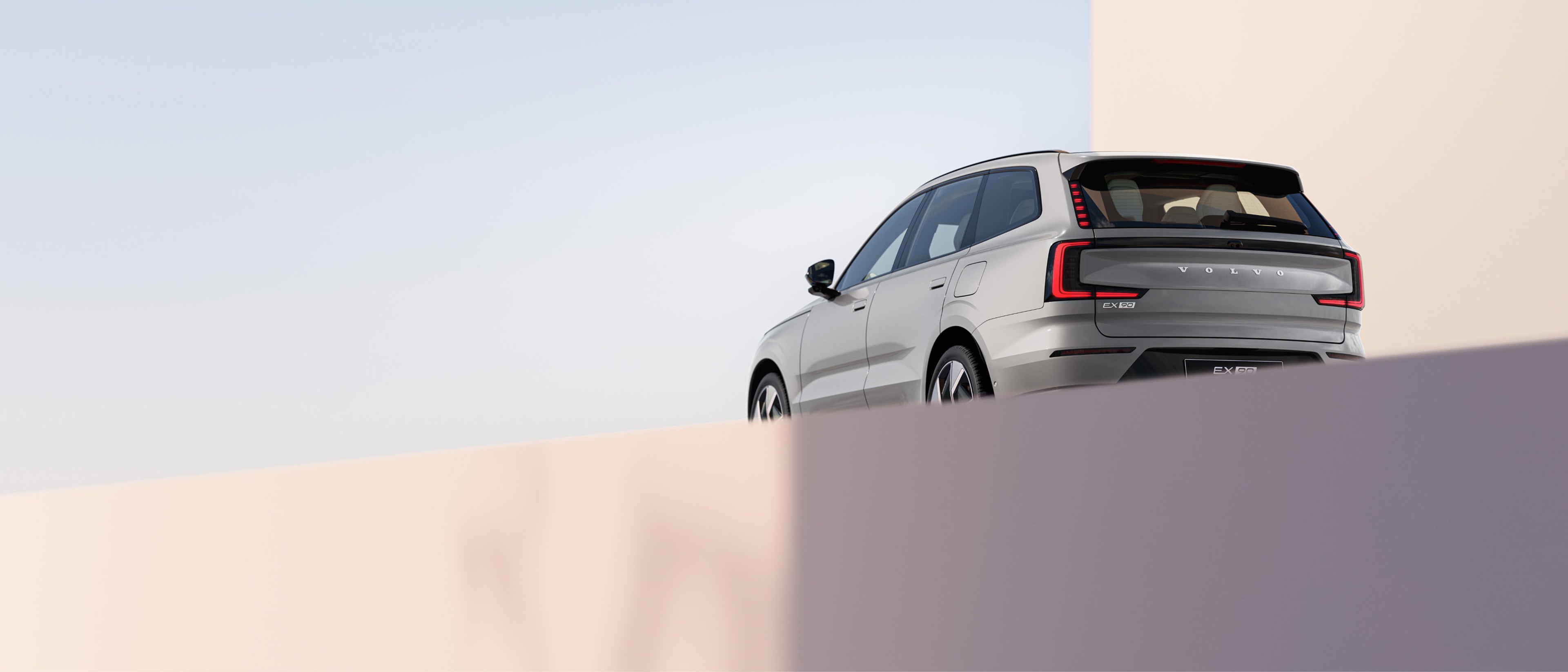 Rear view of a silver Volvo EX90 SUV parked near minimalist architecture in an outdoor space.