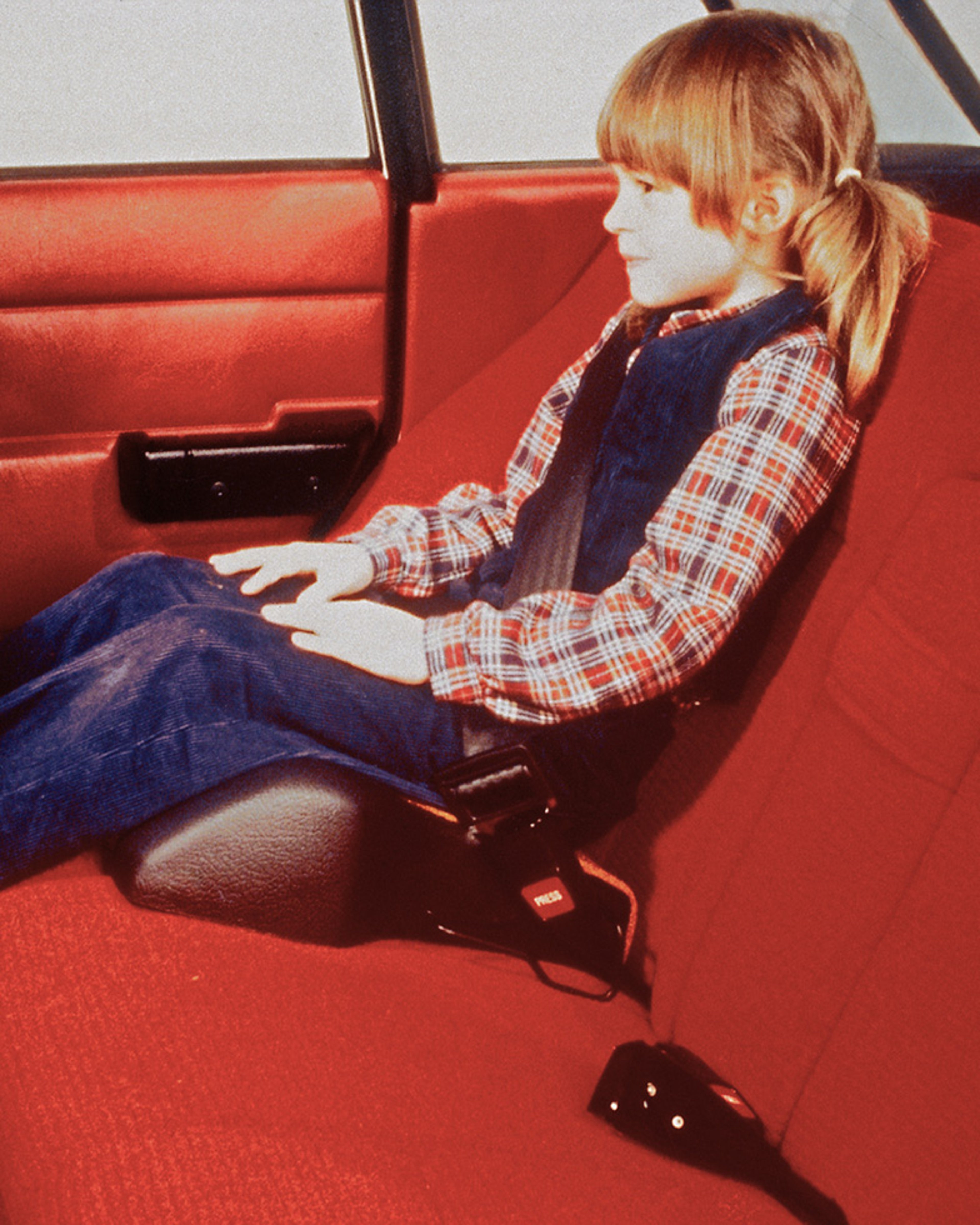 A child seated in the back of a red car interior using a booster cushion and safety belt.