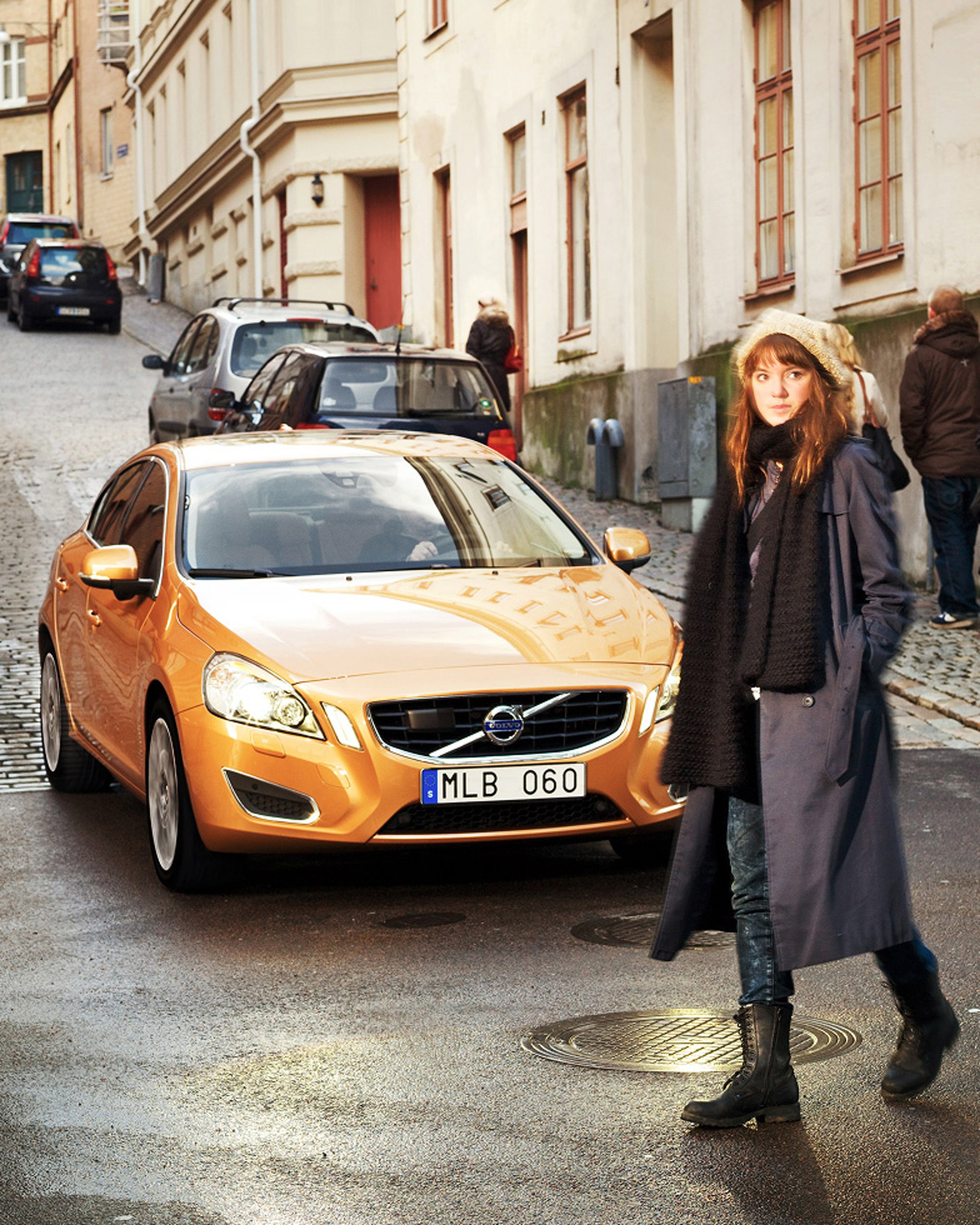An orange Volvo car is stopped in a narrow street while a woman walks in front of it.