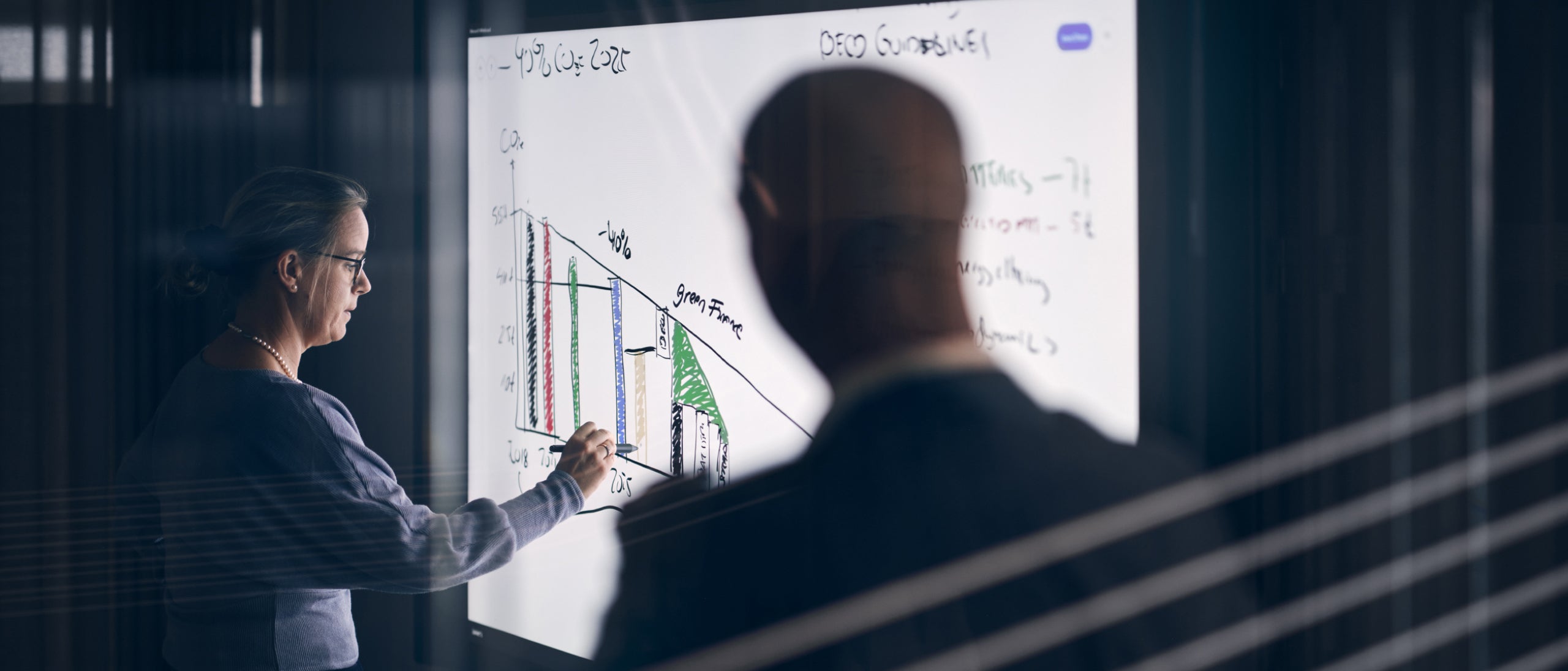 Un homme observant une femme qui travaille sur un tableau intelligent avec un stylet.