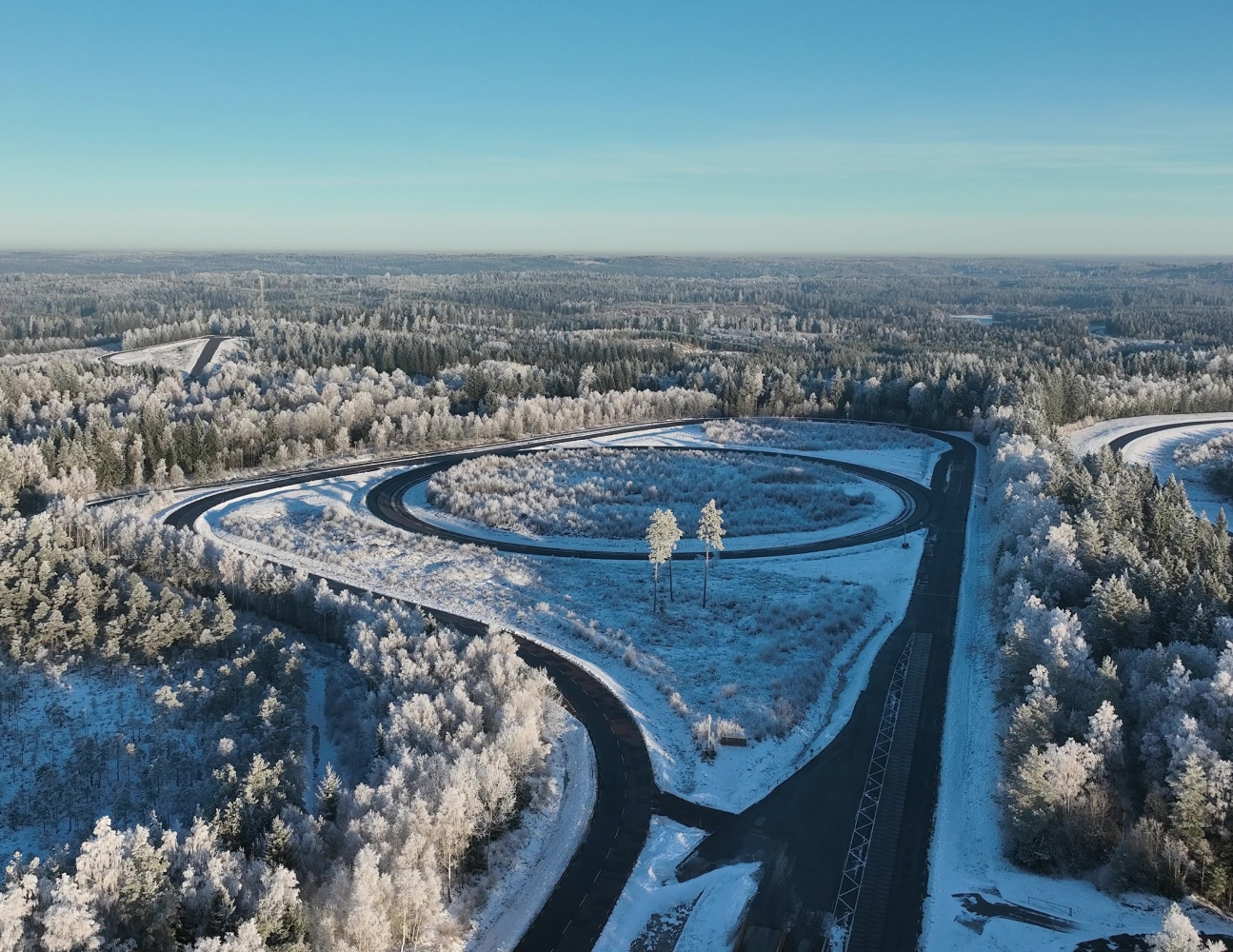 Flygfoto över en snötäckt testbana som slingrar sig genom ett frostigt skogslandskap under en klarblå himmel.