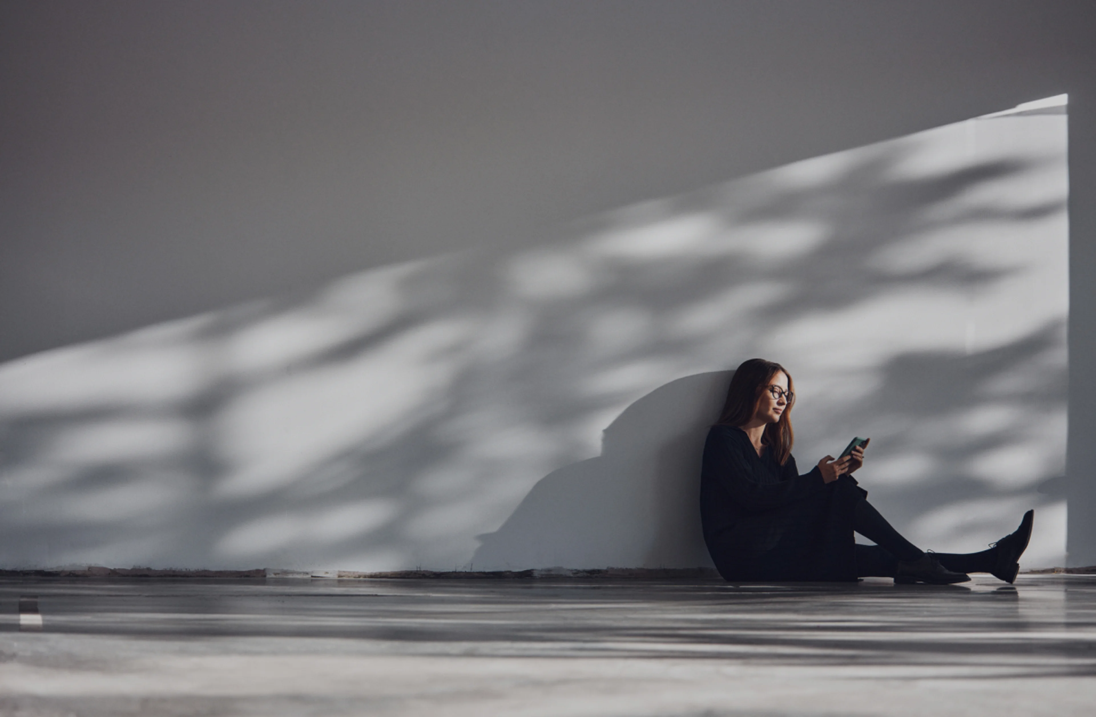 A lady sat on the floor, leaning against a grey wall browsing her phone.