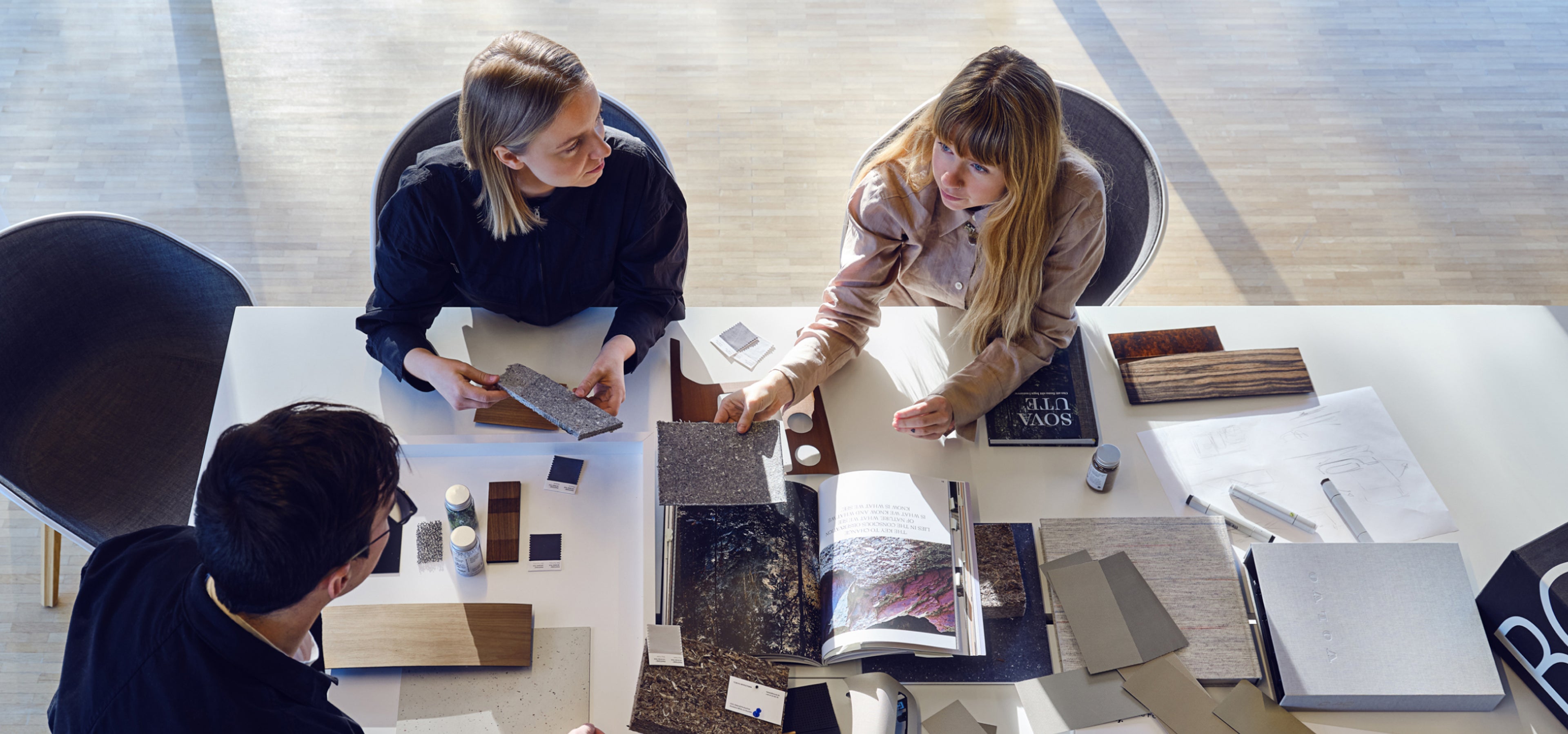 兩個女子和一個男子圍坐在一張桌子旁,討論桌上的材料。