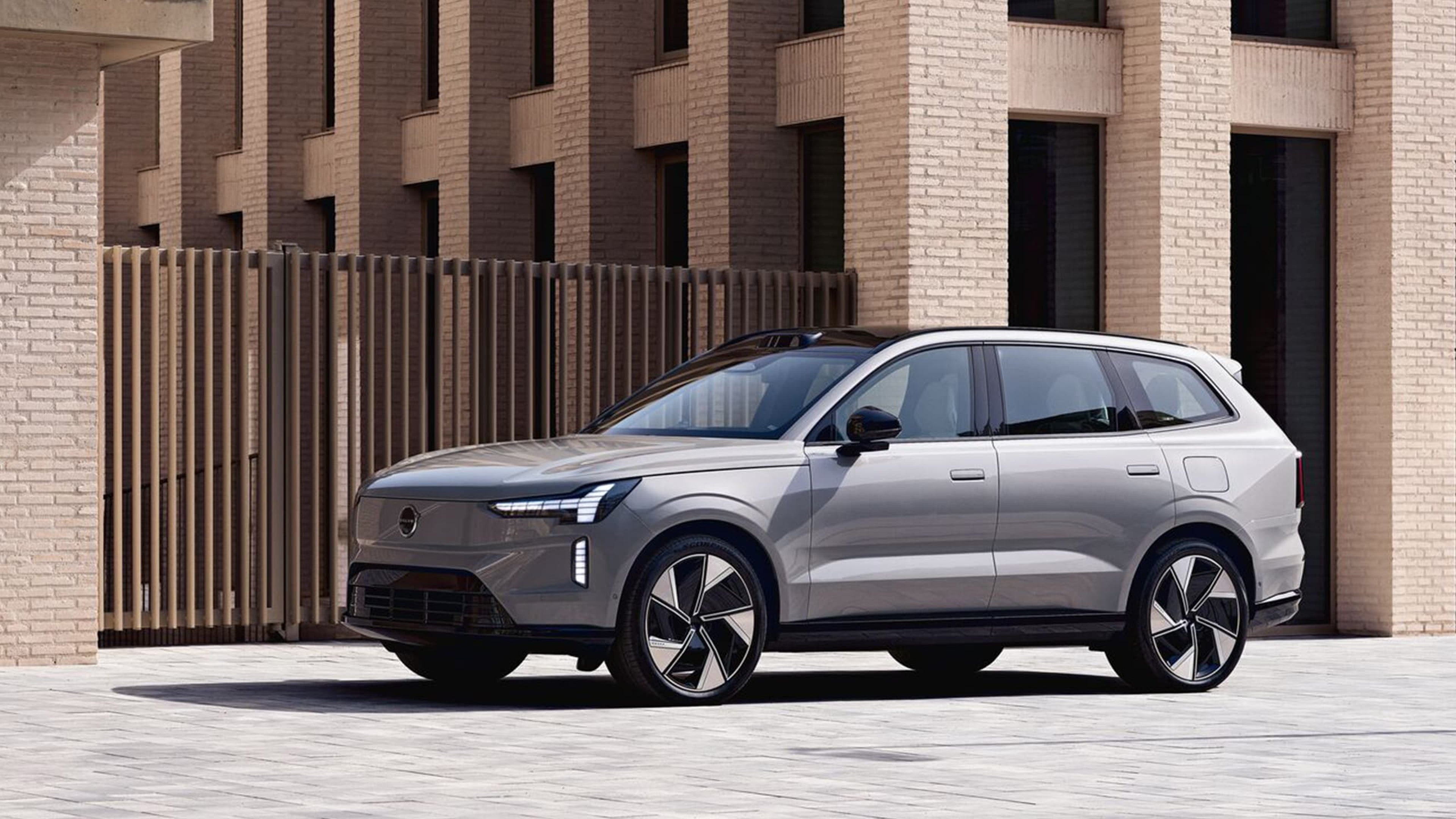 Vue de trois quarts avant de la Volvo EX90 argentée, garée devant un bâtiment moderne avec une façade en briques.