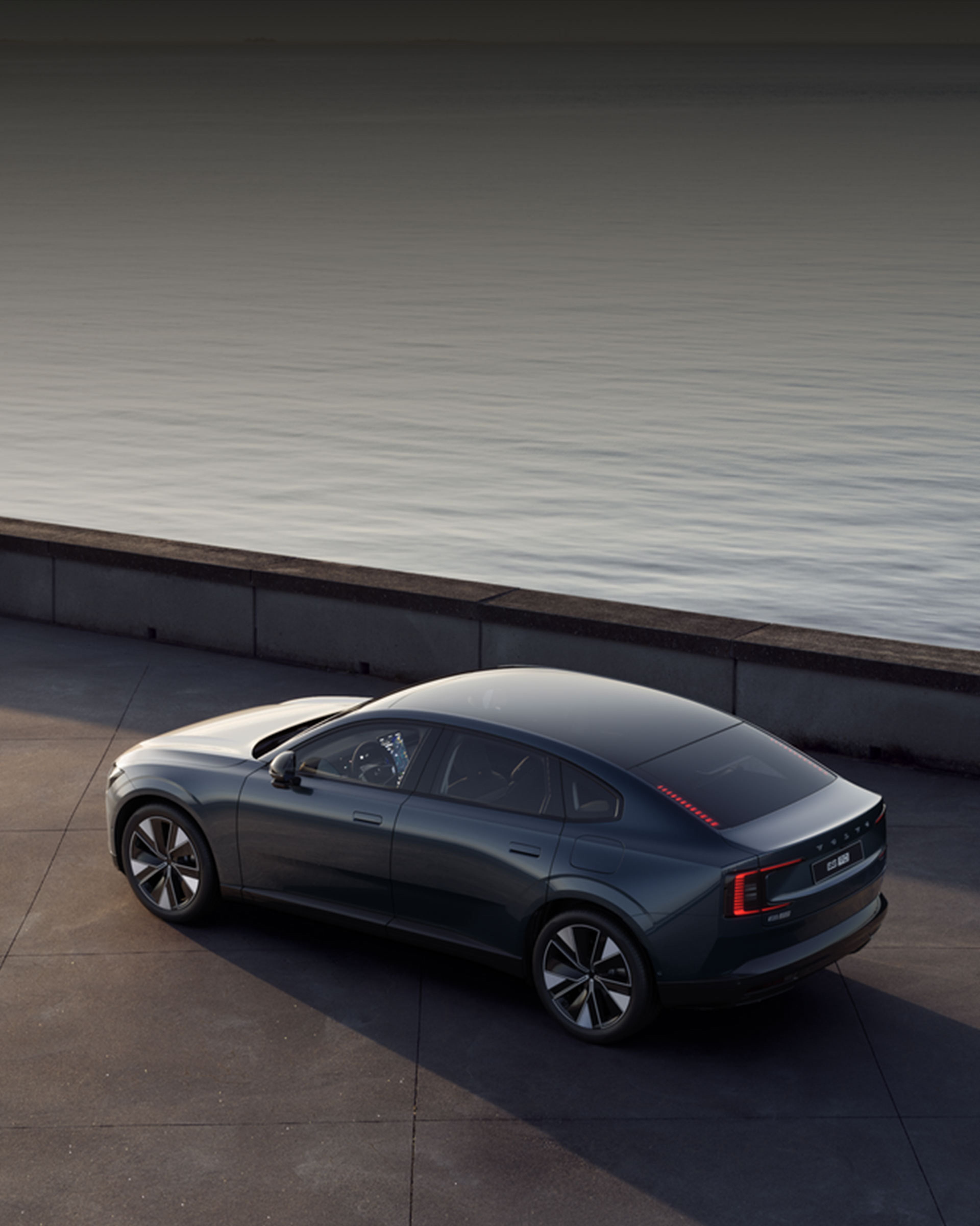 Berline électrique sombre garée près d’un muret, vue arrière, avec la mer en arrière-plan sous ciel gris.