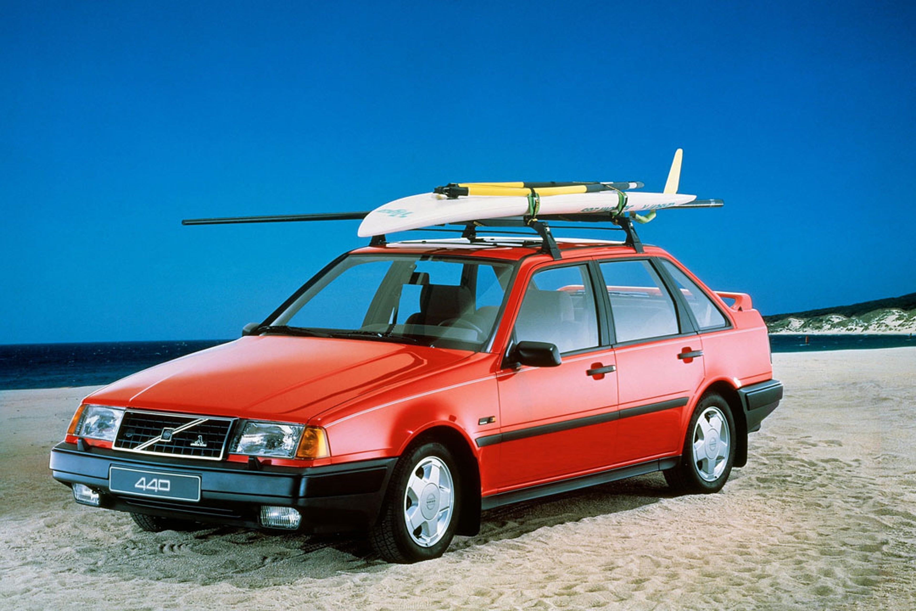 Un Volvo 440 aparcado en la playa con una tabla de surf en la baca.