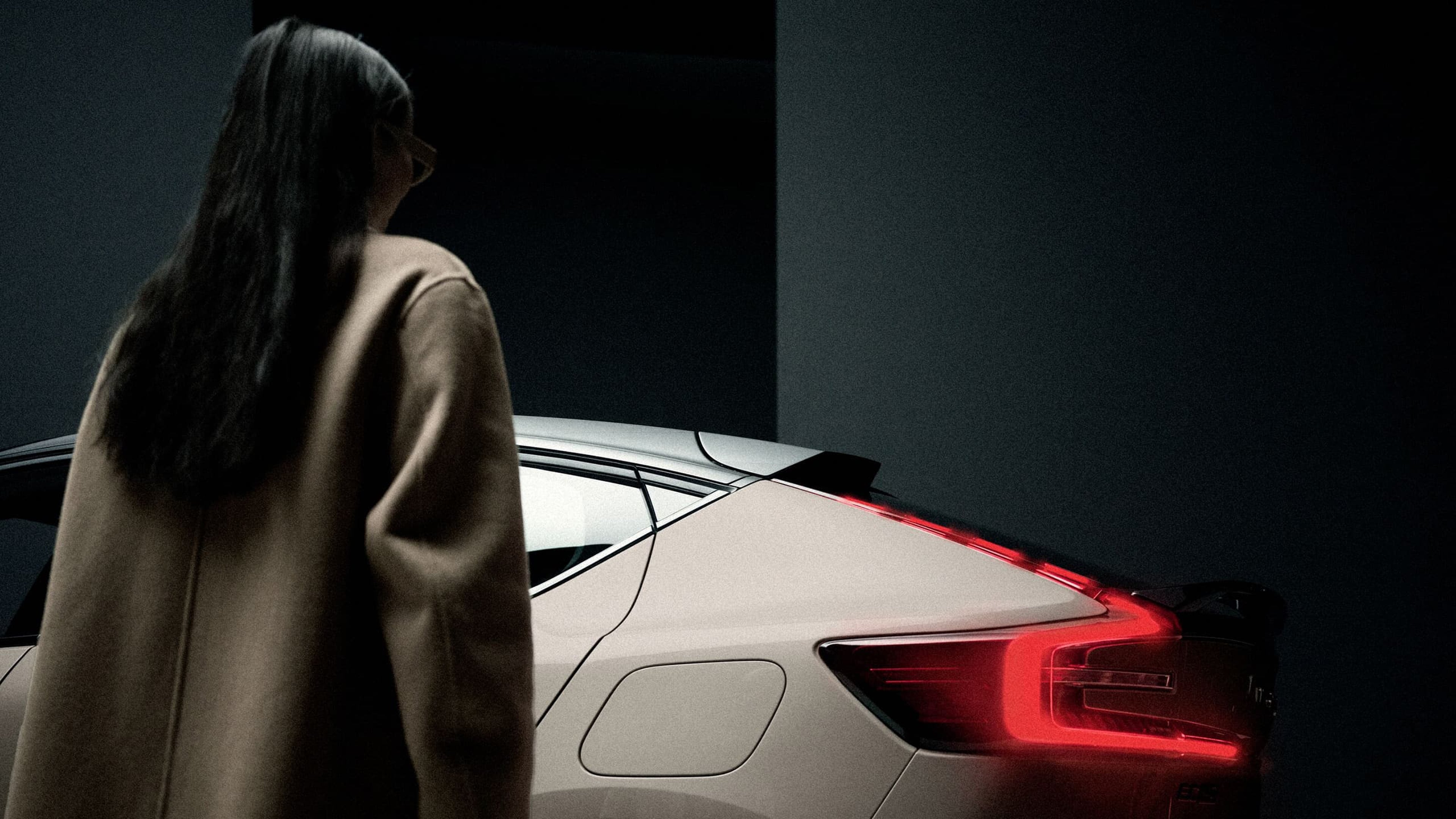 Een vrouw, van achteren gezien, die naar de achterkant van een Volvo EC40 kijkt, waarbij de kenmerkende LED-achterlichten oplichten in een donkere omgeving.