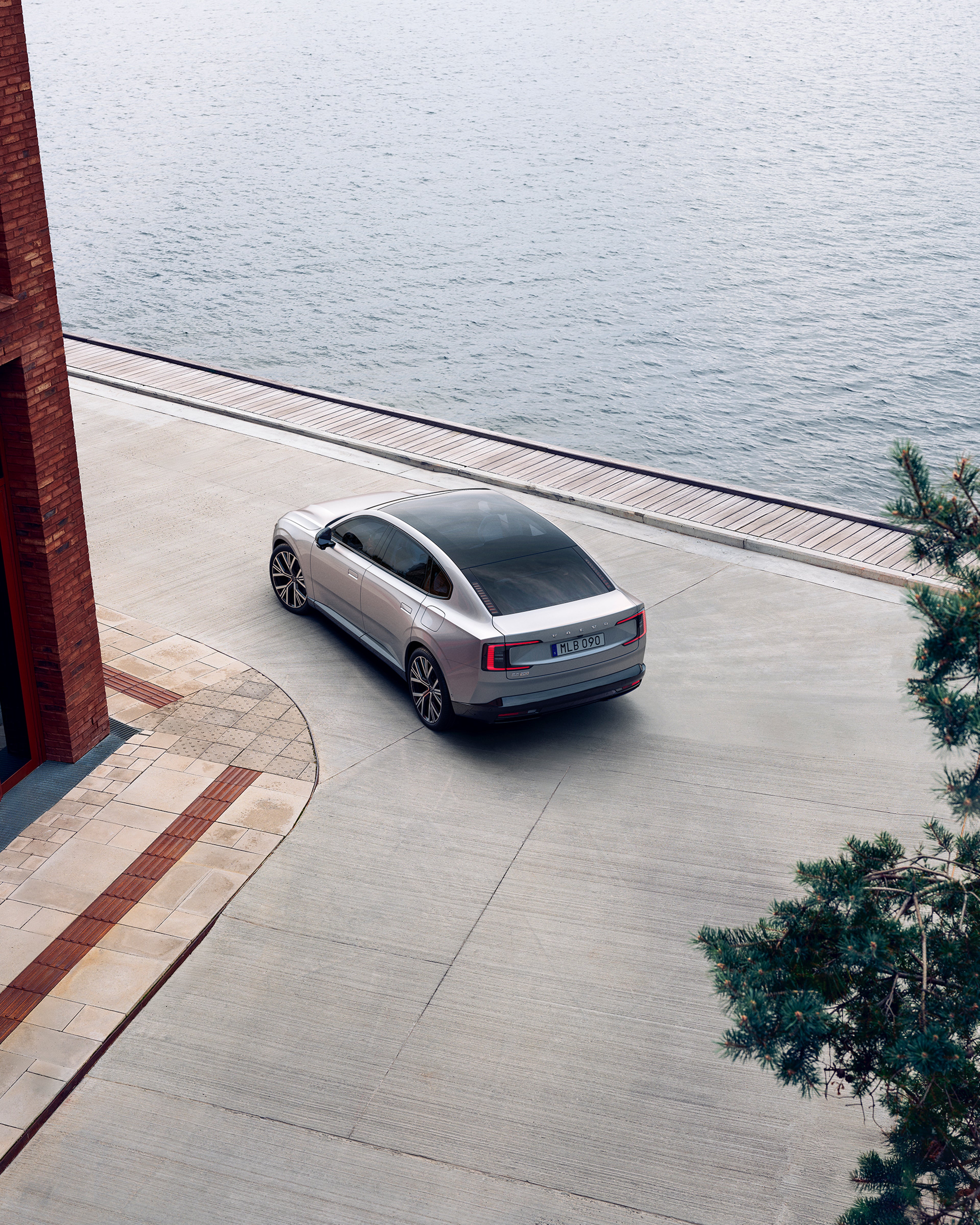 Skats no aizmugures uz Volvo ES90, kas brauc gar krastmalu netālu no ķieģeļu ēkas.