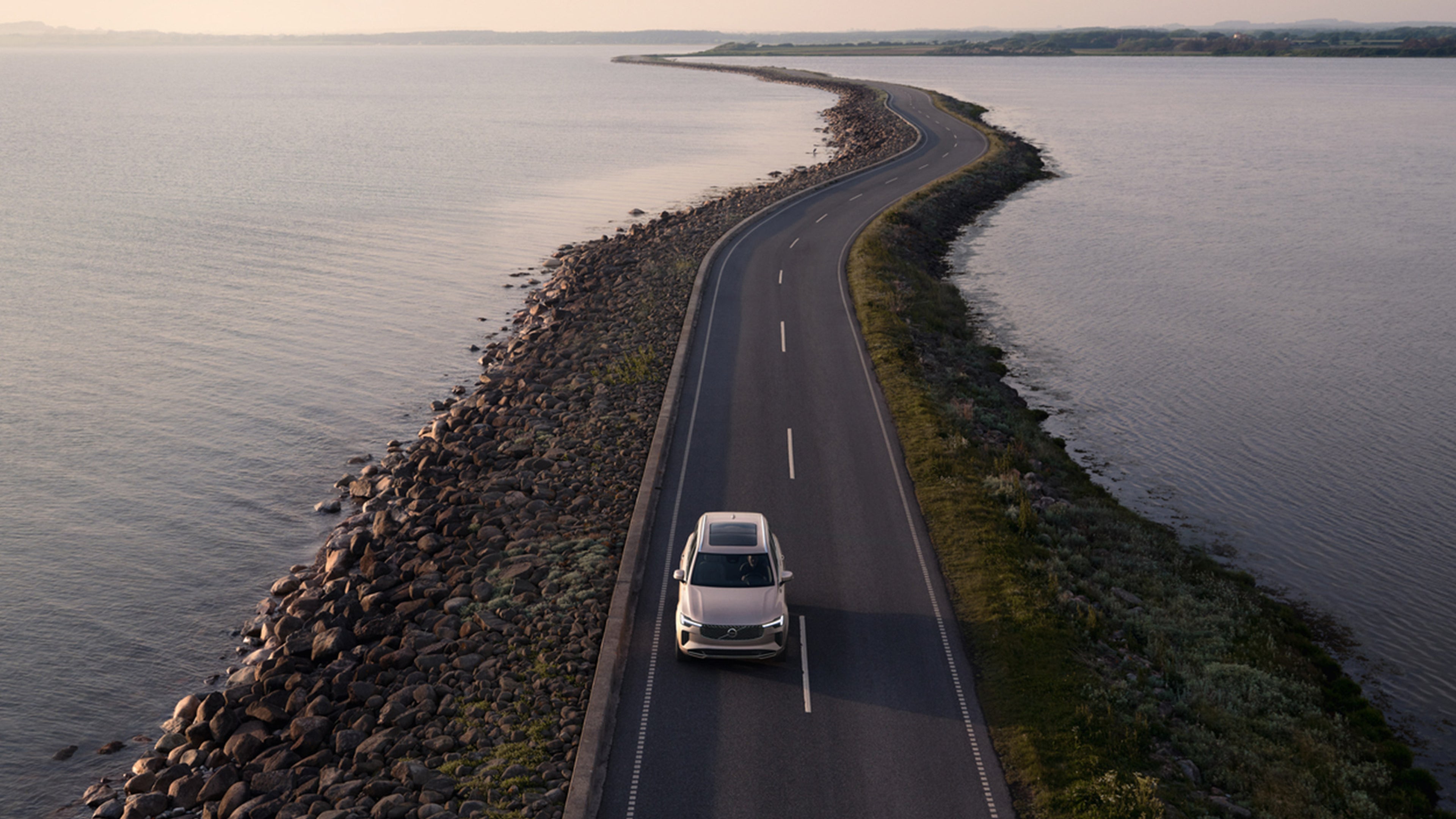 Bright dusk -värinen Volvo XC90 lataushybridikatumaasturi ajaa merialuuen ylittävää kannastietä.