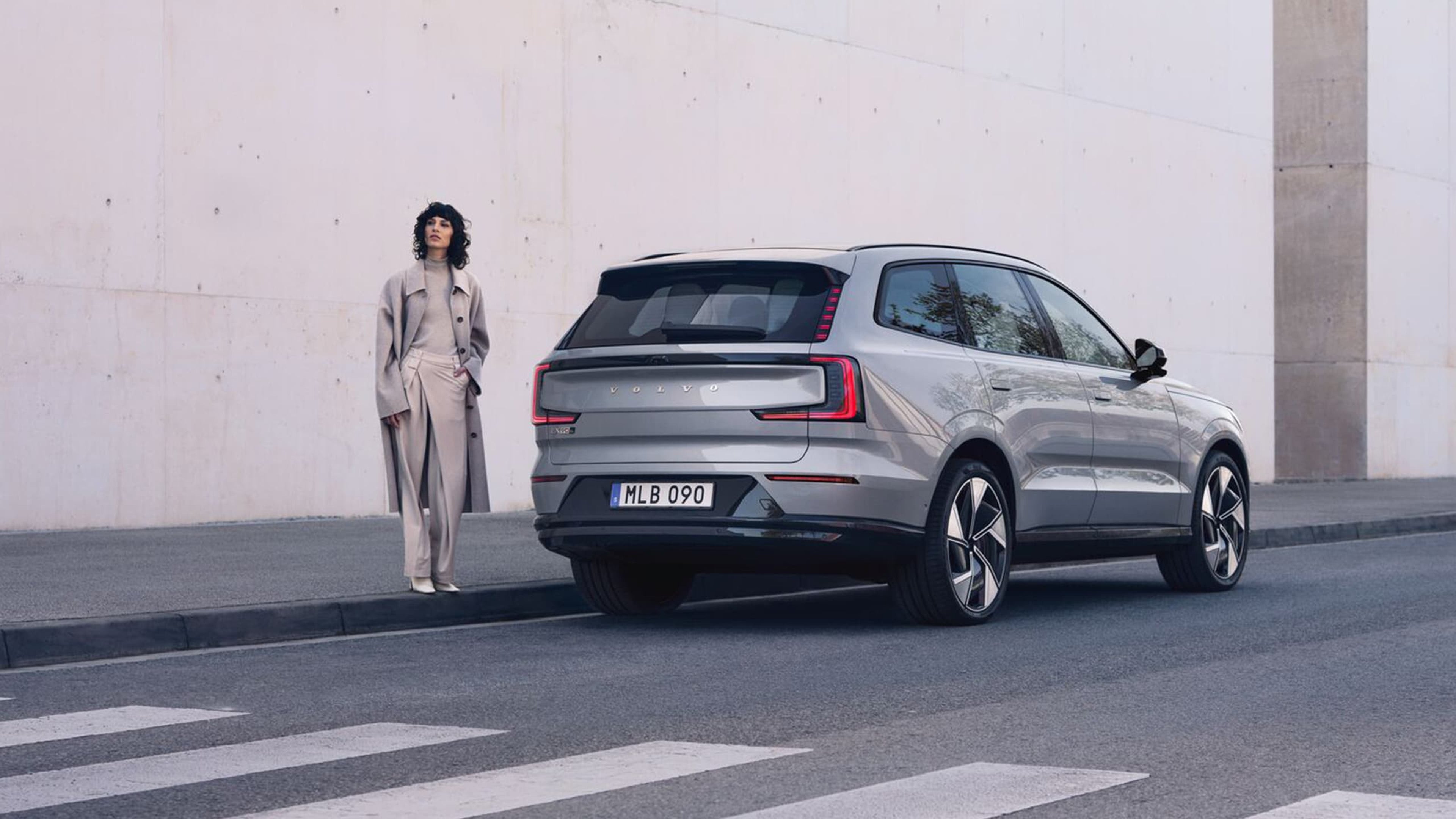 Vue arrière de la Volvo EX90 argentée, garée au bord de la route. Une femme se tient sur le trottoir à côté de la voiture, près d'un passage piéton.